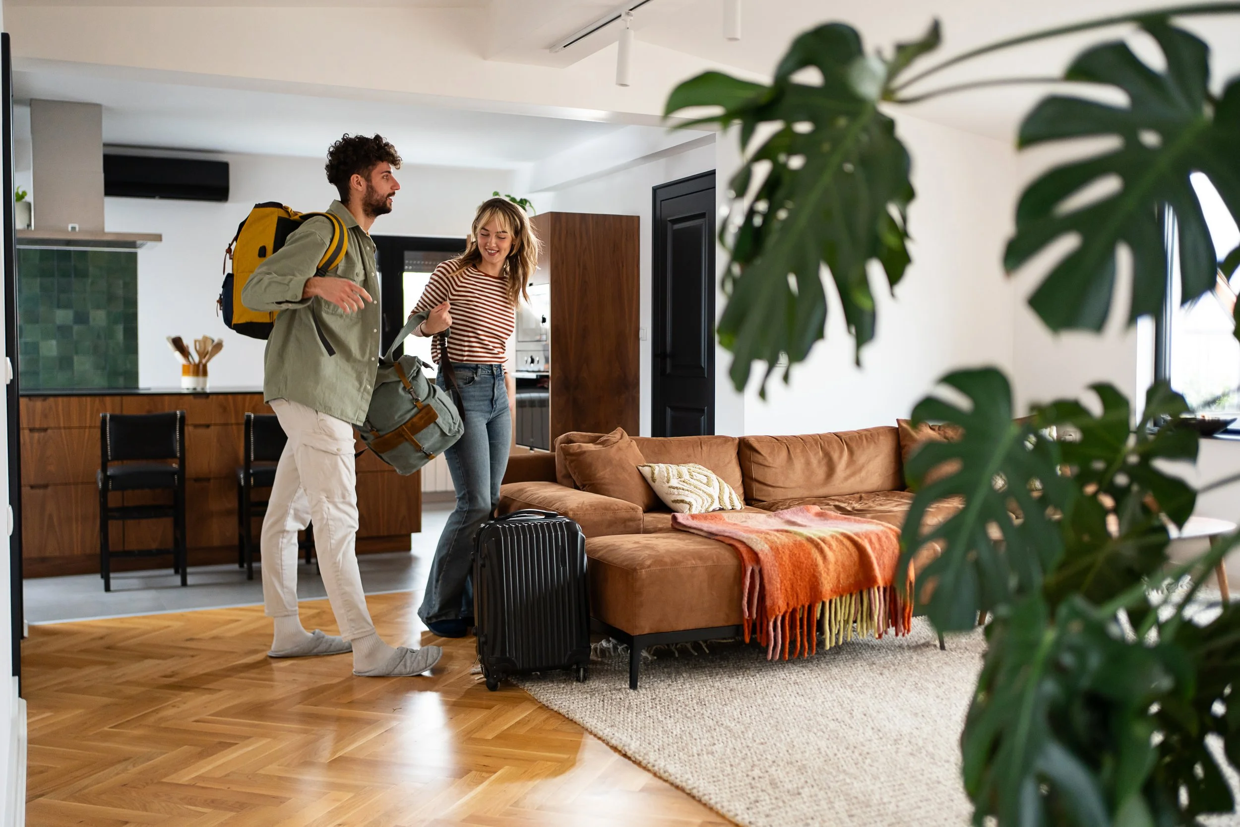 Two people with luggage arriving at a modern apartment, smiling and greeting each other, with a kitchen and living room visible in the background.