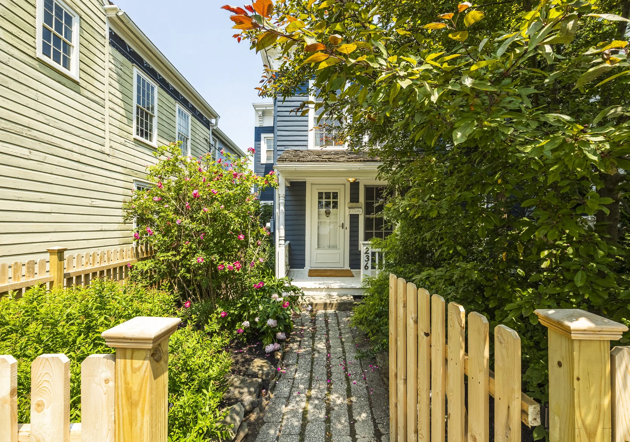 Front entrance of a house with a white door, surrounded by green foliage and pink flowers, with a wooden fence and a pathway leading up to the porch.