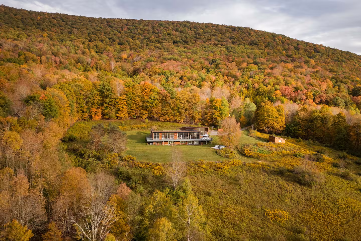 Modern house on a green lawn with a backdrop of a forested hillside in autumn colors.