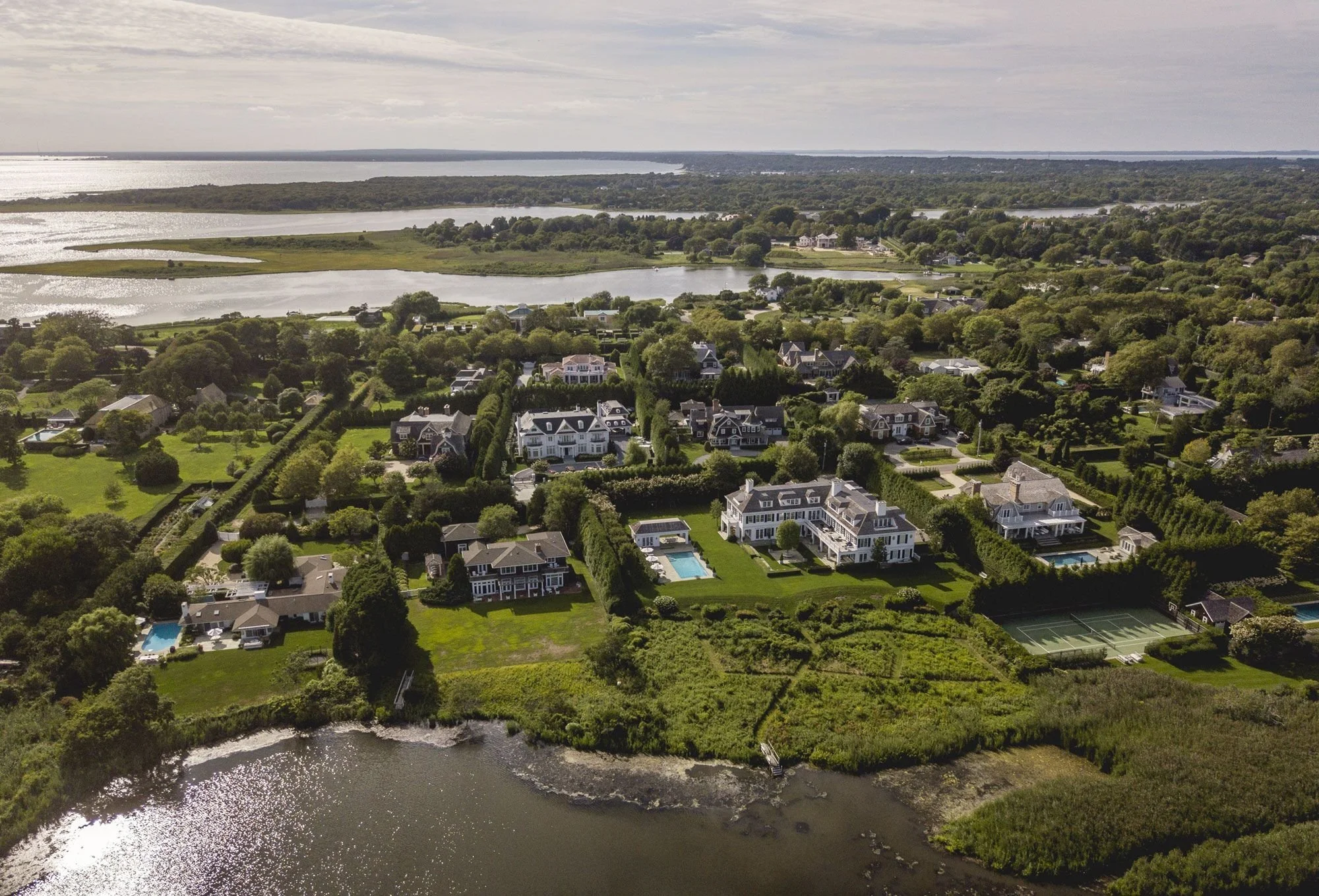 Aerial view of a coastal residential neighborhood with large houses, swimming pools, tennis courts, and green lawns near water bodies and marshlands.