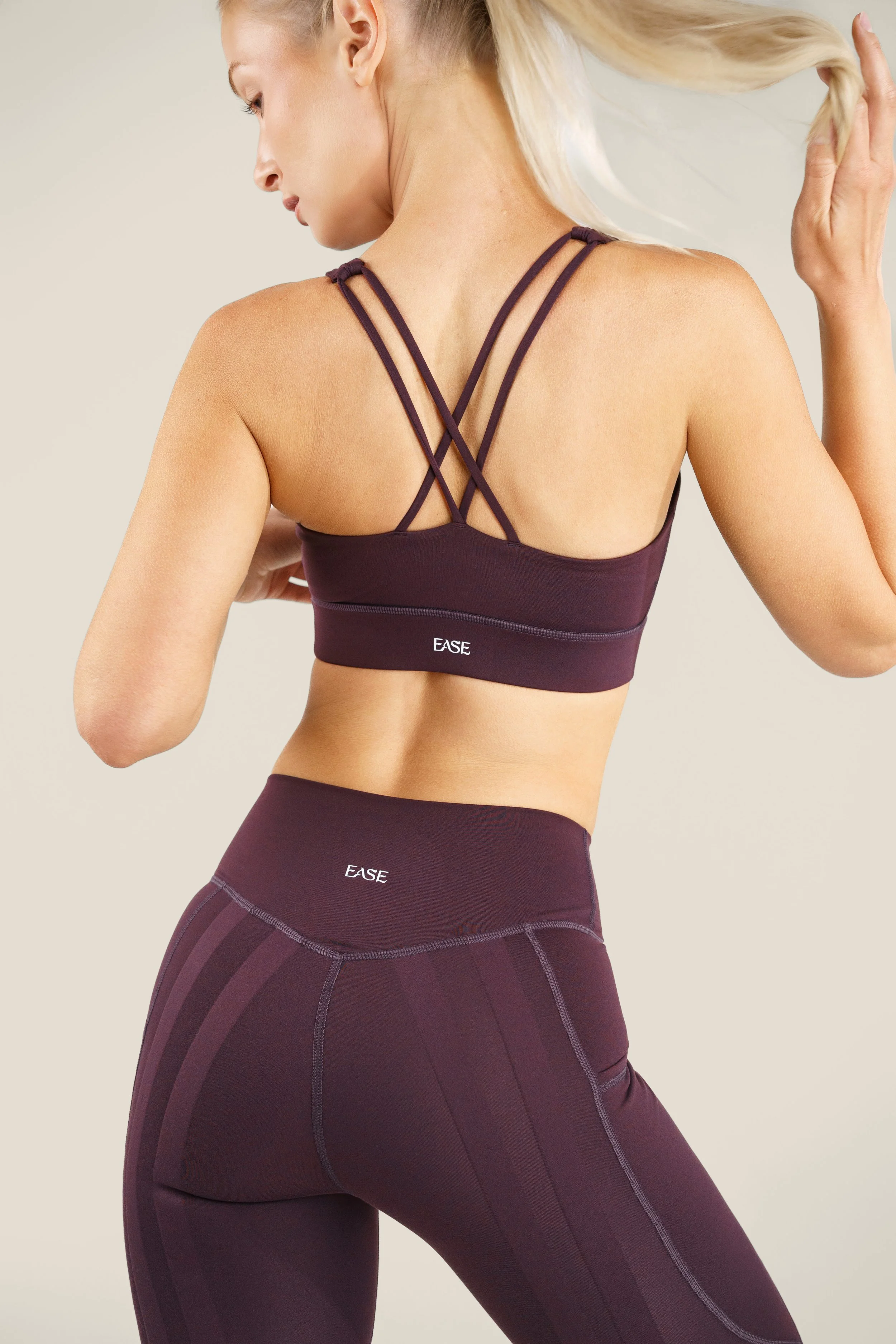 A woman in a purple sports bra and matching leggings, with the word 'EASE' printed on both, is shown from the back, fixing her blonde hair with her right hand.