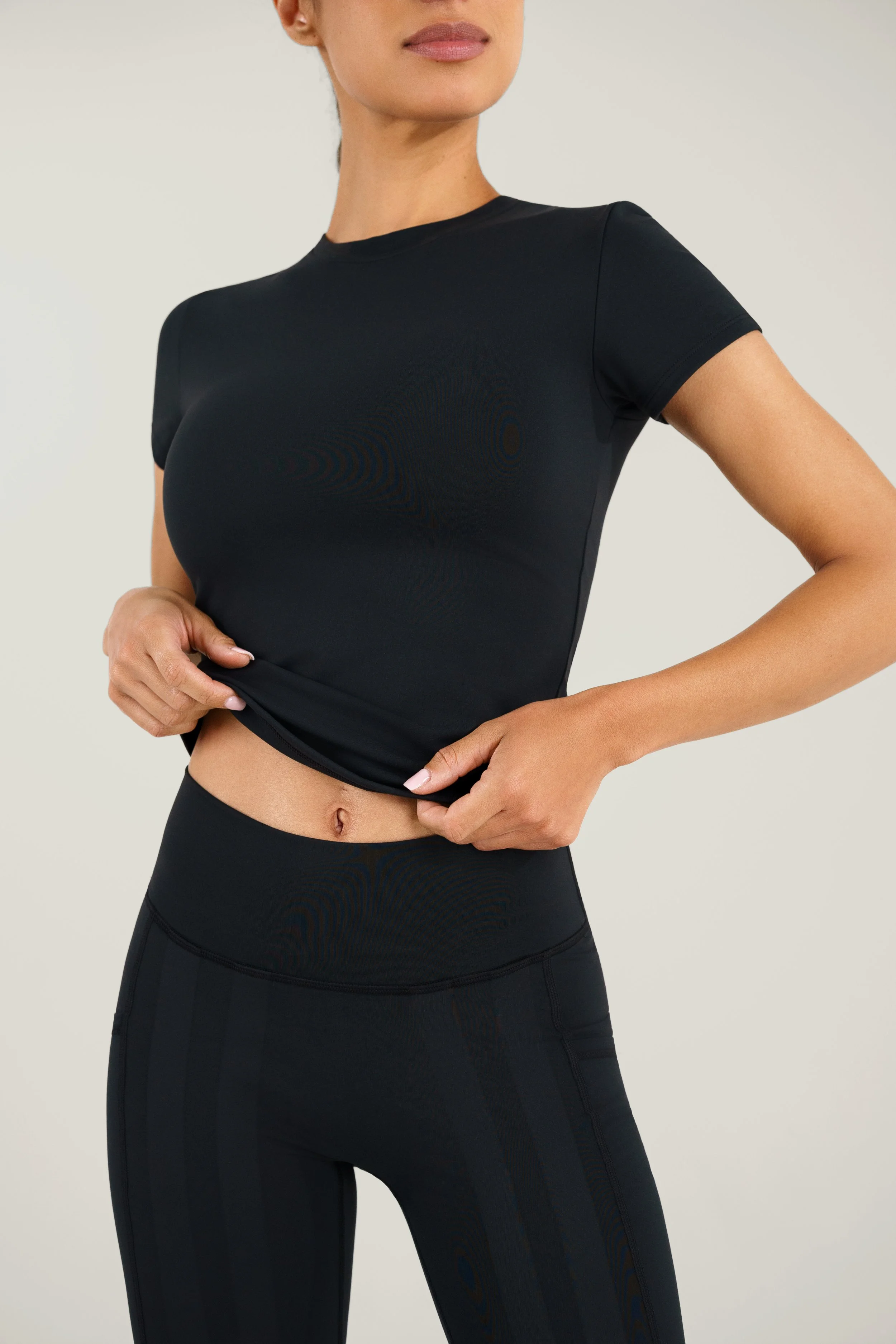 A woman is wearing a black fitted athletic t-shirt and matching black high-waisted leggings, standing against a plain background, with her hand lifting the bottom of her shirt.