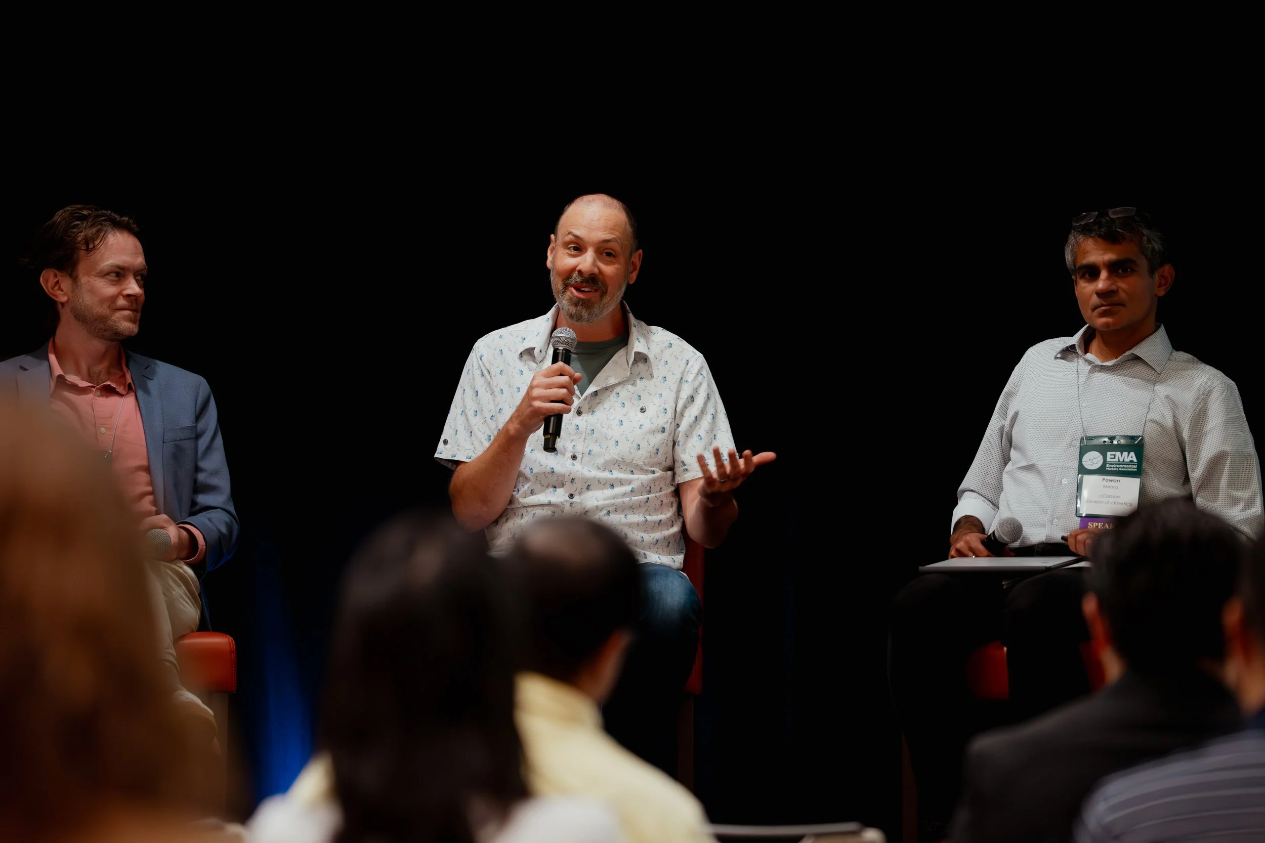 Three men sitting on a stage during a panel discussion, one speaking into a microphone, with audience members in the foreground.