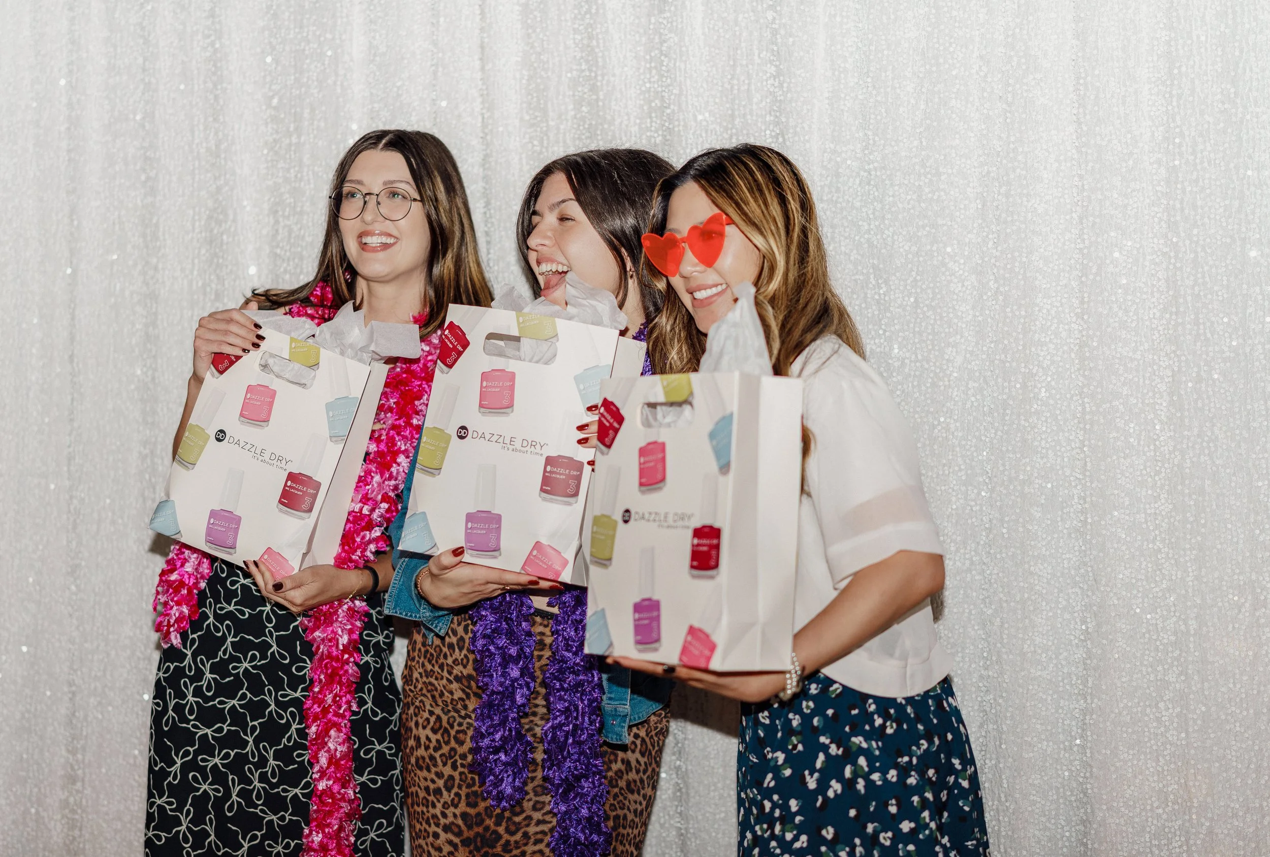 Three women celebrating with gift bags and accessories, dressed casually, in front of a glittery silver background.