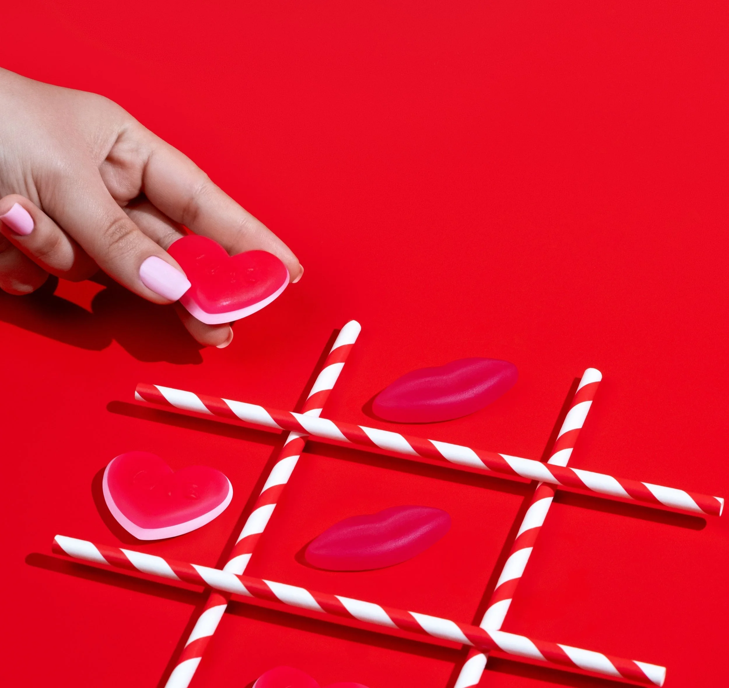 Close-up of a hand holding a pink heart-shaped object with red and white striped straws scattered on a red background.