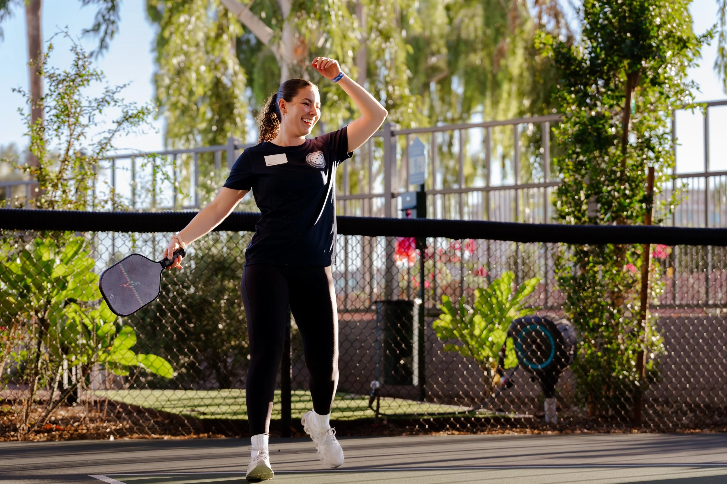 A woman smiling and dancing on an outdoor tennis court, holding a paddle in her right hand, surrounded by trees and sunlight.