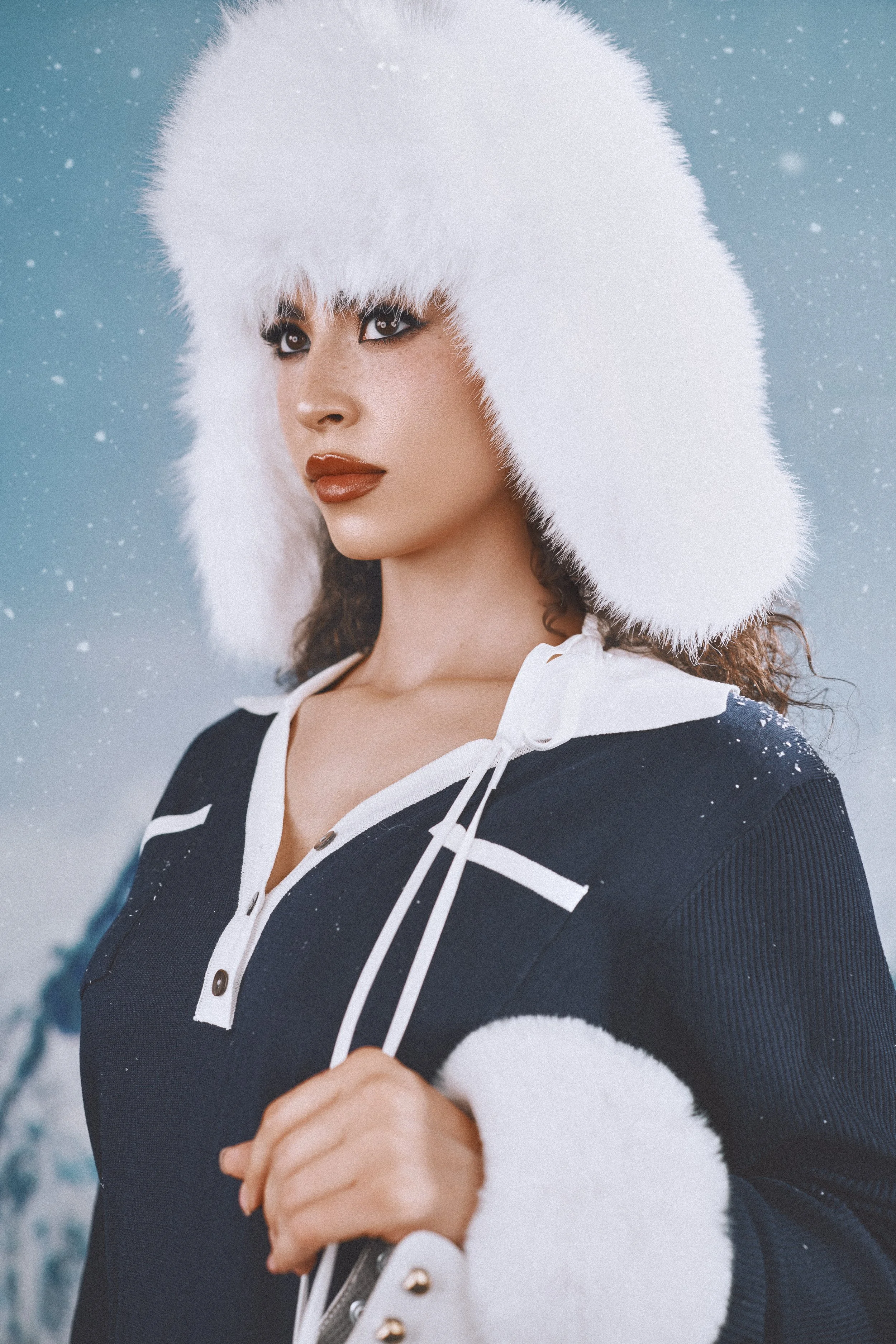 A woman wearing a white fluffy winter hat and a navy blue sweater with white accents, standing outdoors in snowy weather.