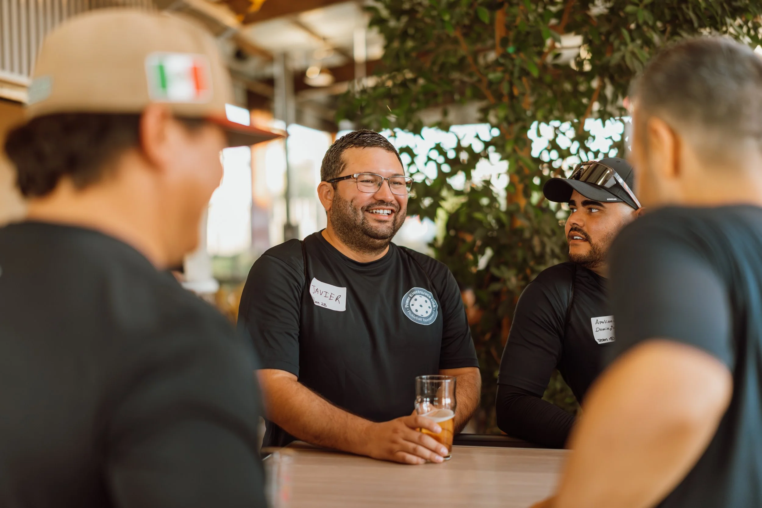 Four men sitting and talking at a table outdoors, with one man smiling and holding a glass of beer; three men are partially visible, and in the background there are trees and a building.