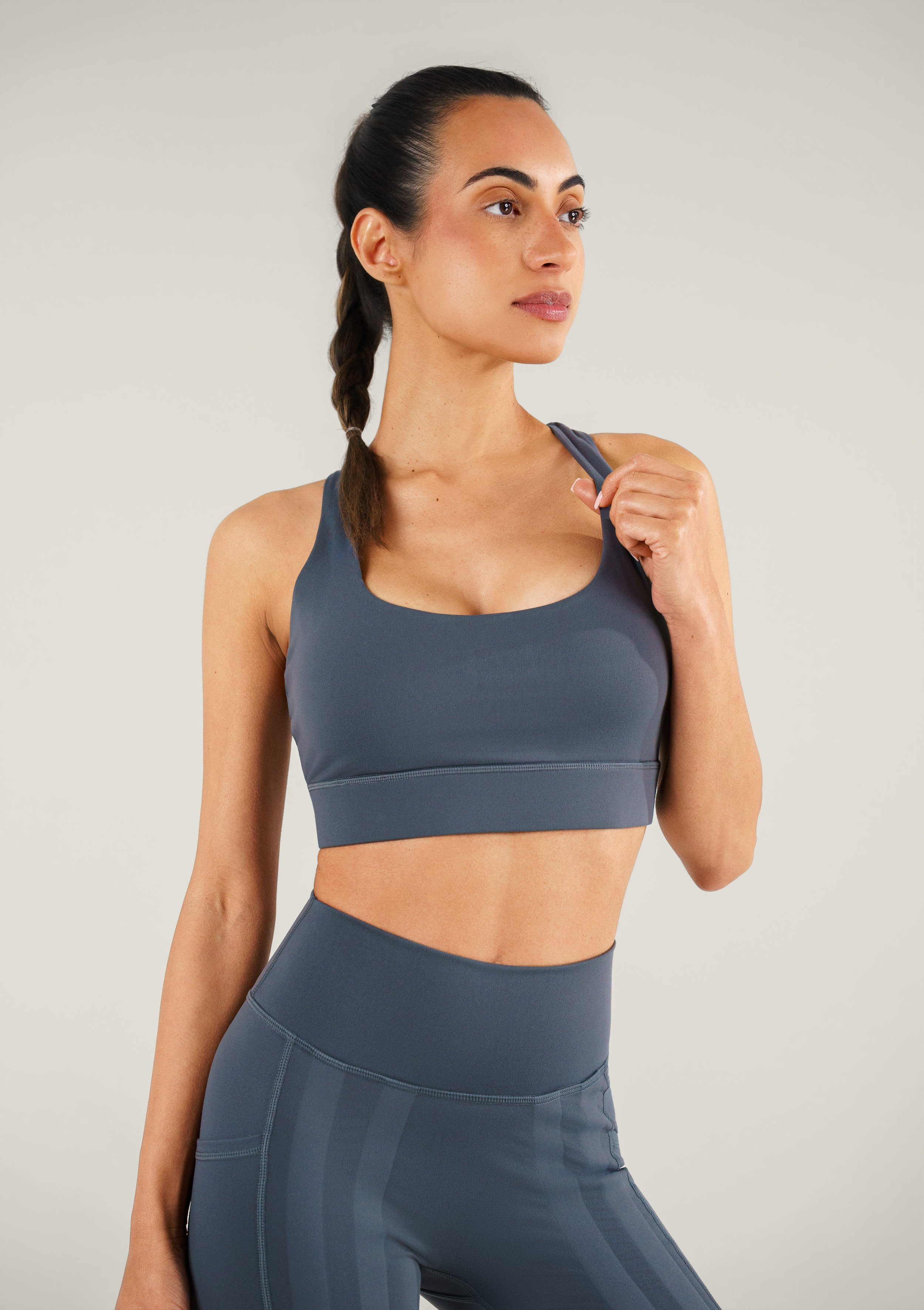 A woman with dark hair in a braid wearing a gray sports bra and matching leggings, standing against a plain neutral background.