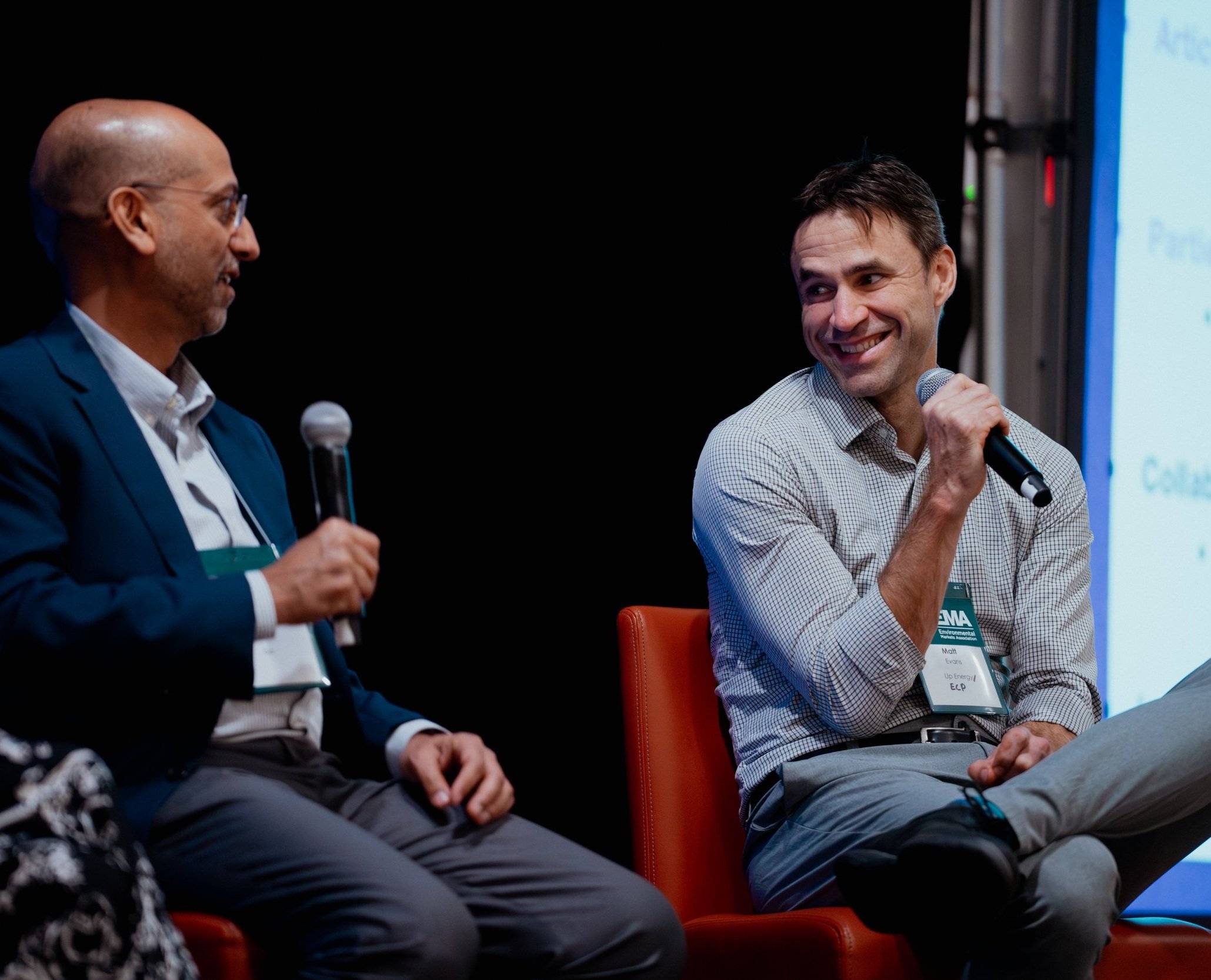Two men sitting on orange chairs holding microphones during a discussion at a conference or event, with a dark background and a presentation screen to the right.
