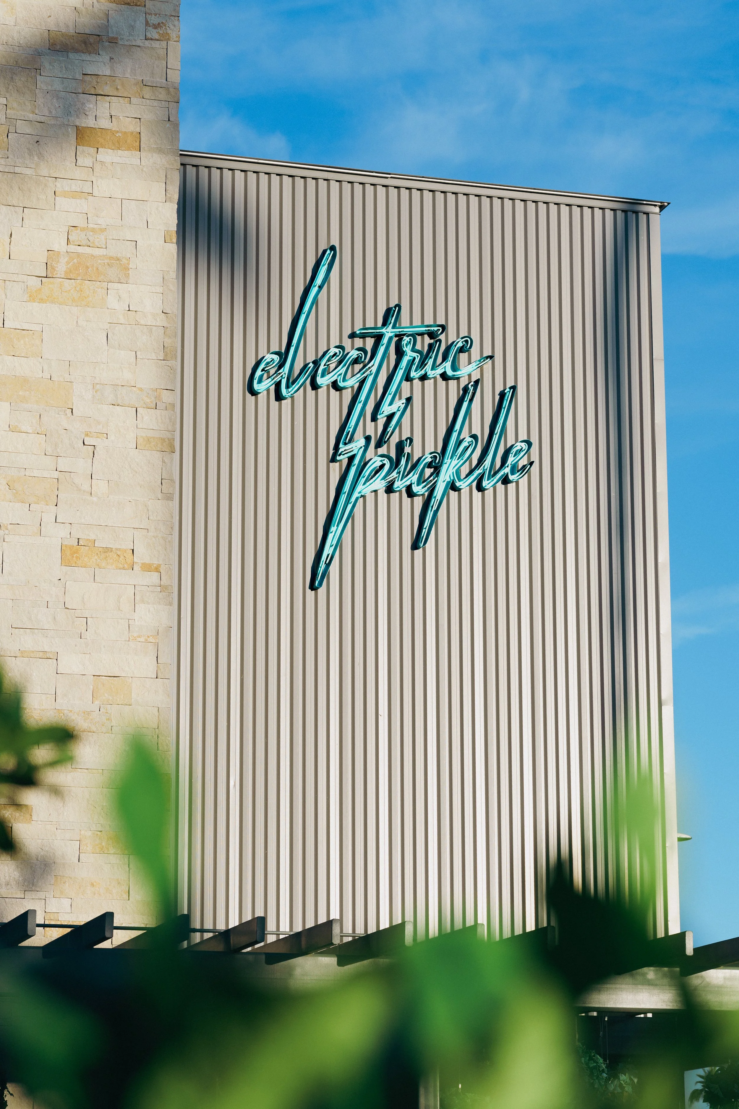 Neon sign reading 'electric twelve' on the exterior of a building with beige stone and metal paneling, under a blue sky.