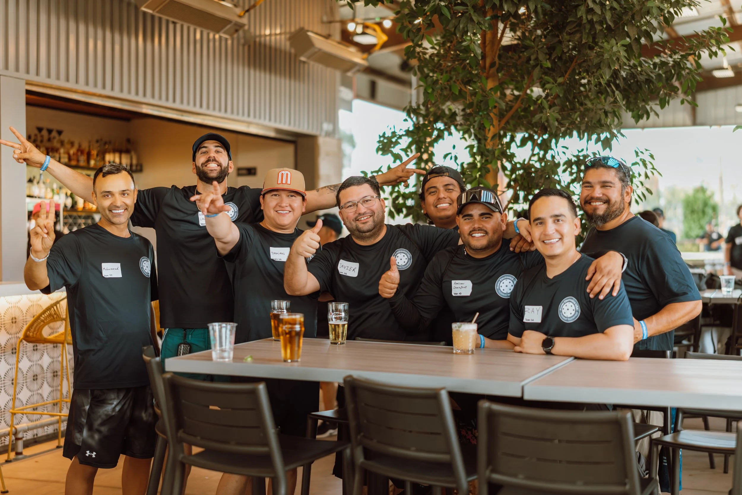 Group of smiling men posing with thumbs up and peace signs at a table in a restaurant or bar, with drinks on the table and a large plant in the background.