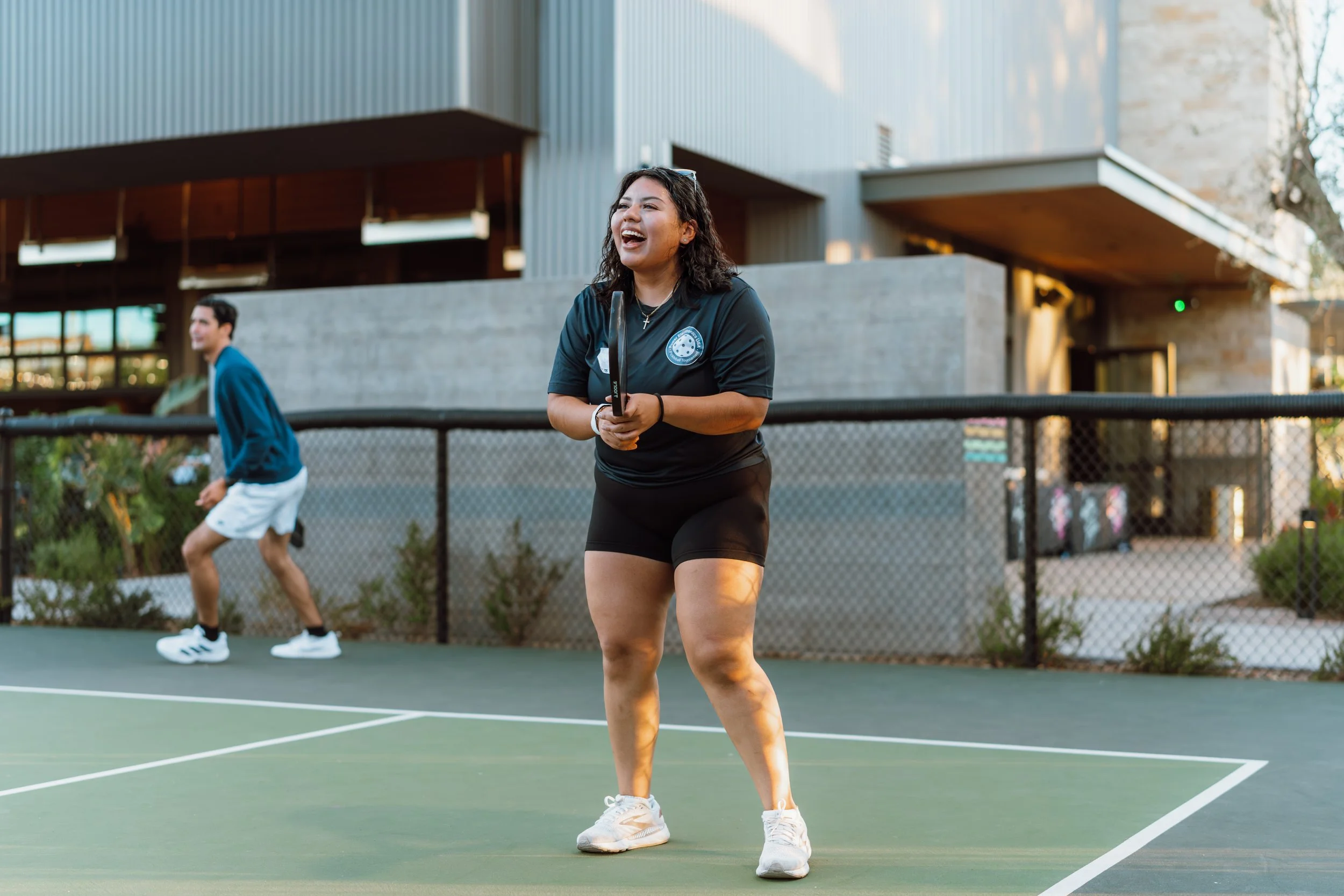 A young woman with curly dark hair playing pickleball on an outdoor court, smiling and holding a paddle, with a young man in the background preparing to hit the ball.