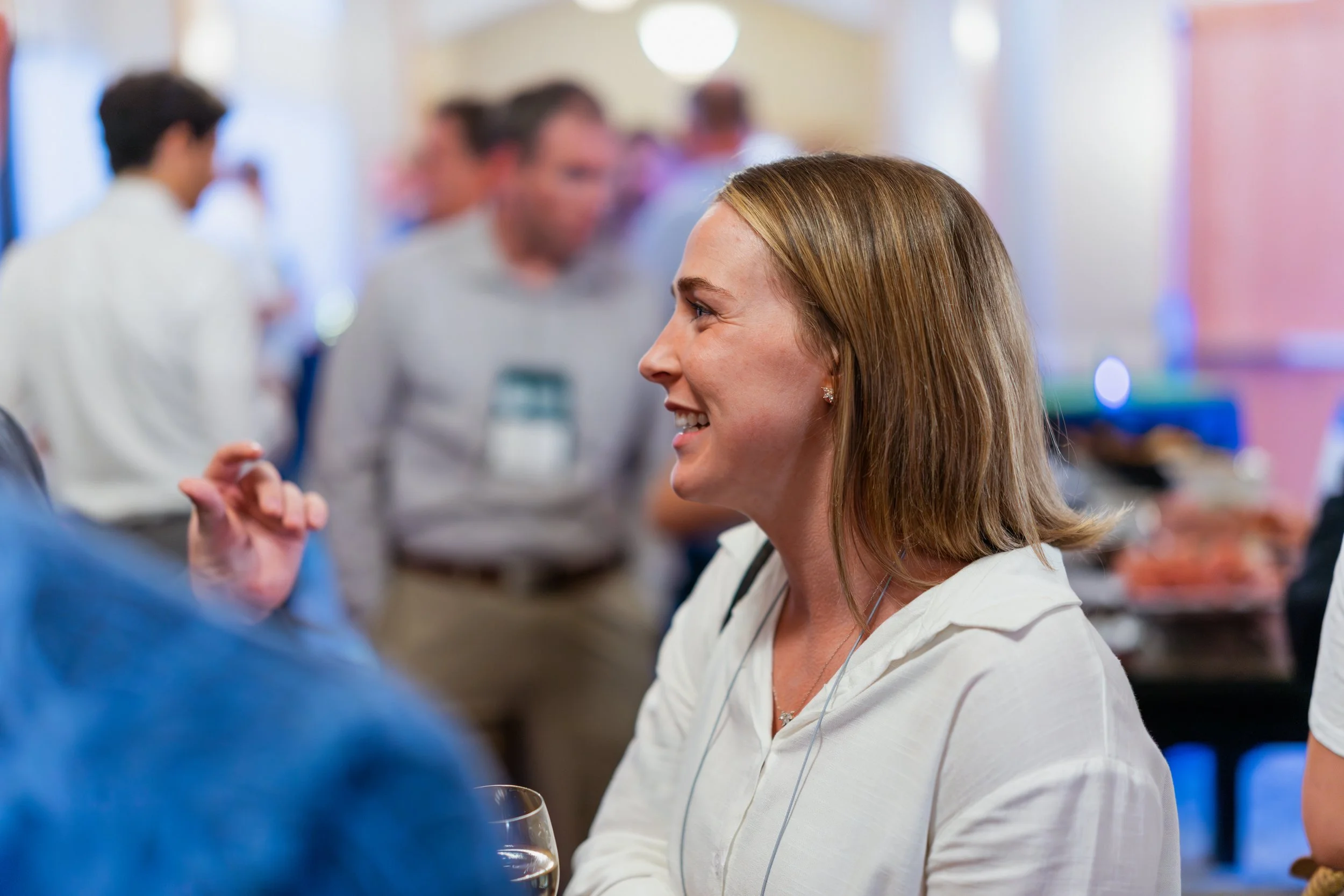 A woman with shoulder-length light brown hair smiling and engaged in conversation at a social gathering, with blurred guests in the background.