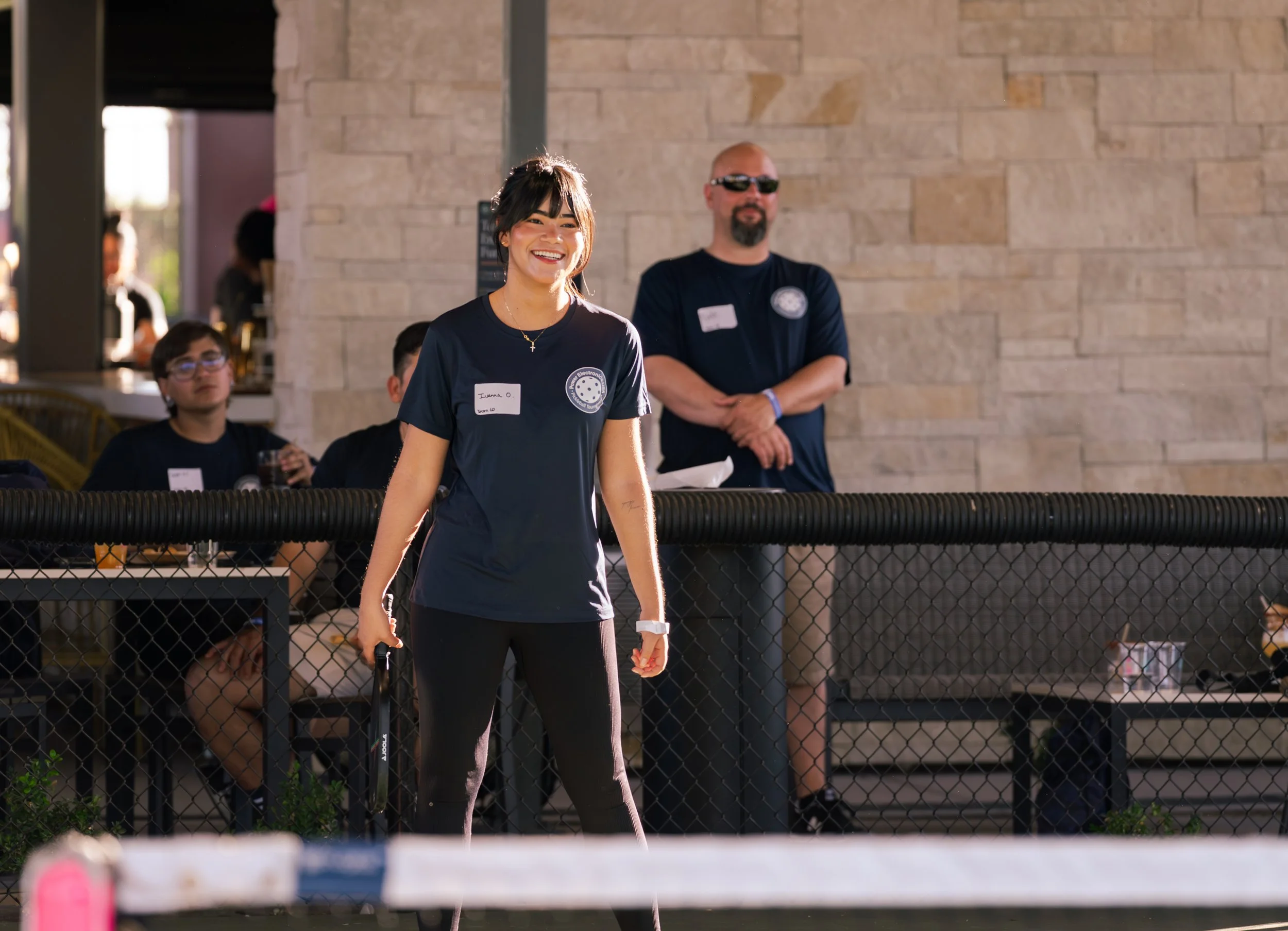 A young woman with dark hair and a bright smile standing outdoors, wearing a navy blue shirt with a logo and a name tag, holding a leash, with a man in sunglasses and others seated behind a black fence in the background.