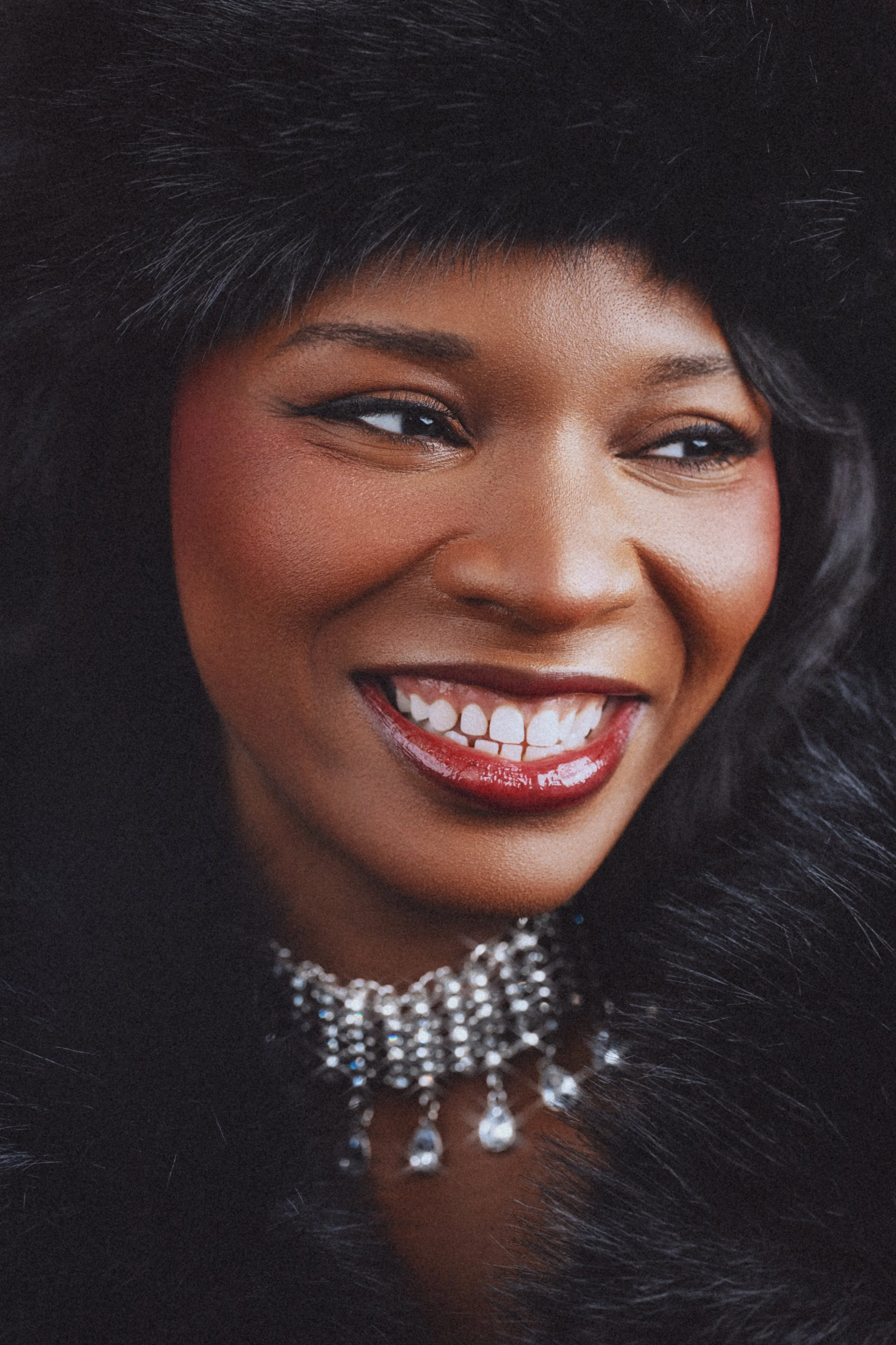 Close-up portrait of a woman smiling, wearing a black fur hat and a sparkling pearl necklace.