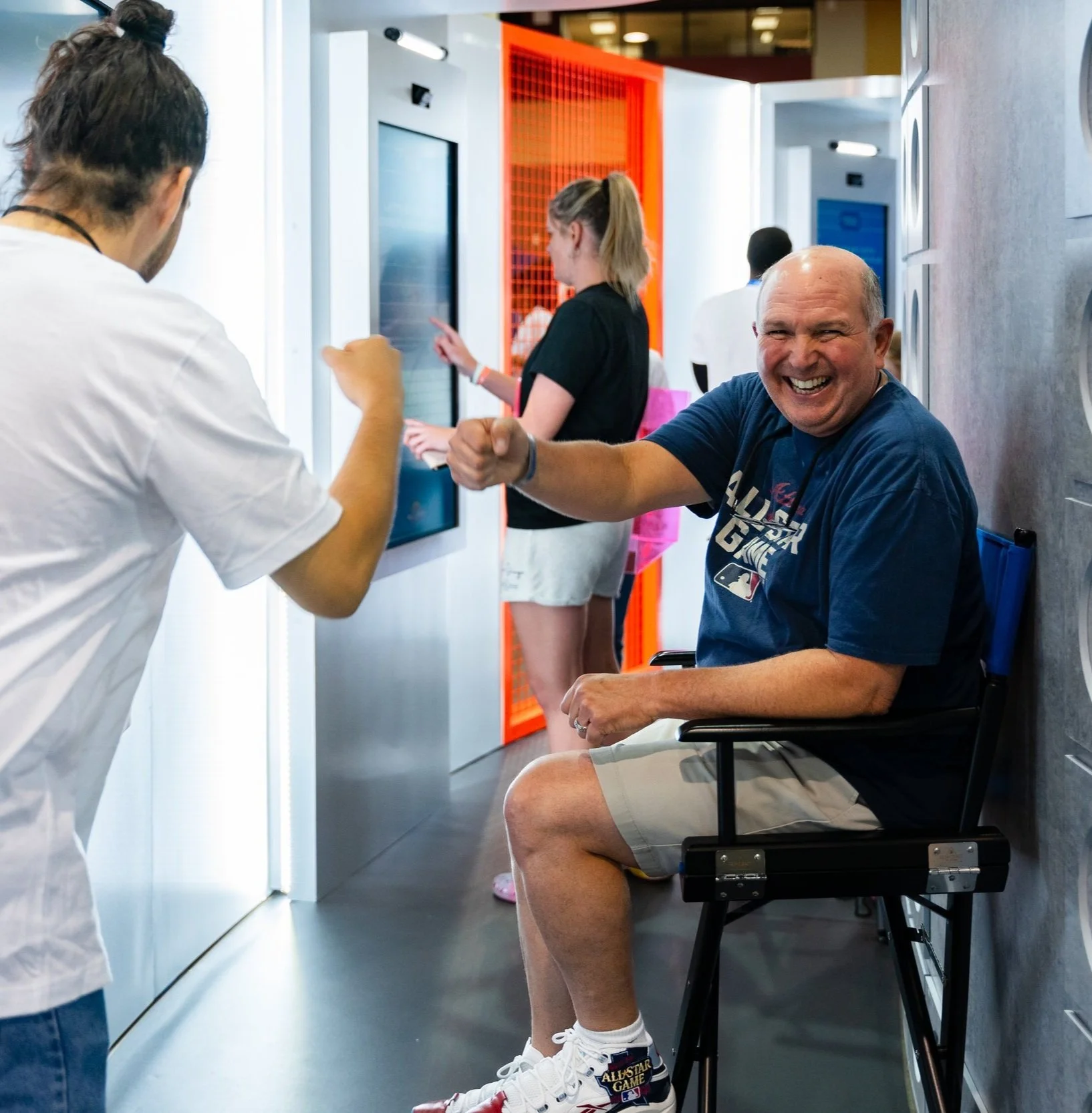A man in a wheelchair smiling and joking with a person wearing a white shirt, with others in the background using touchscreen kiosks in a modern indoor setting.