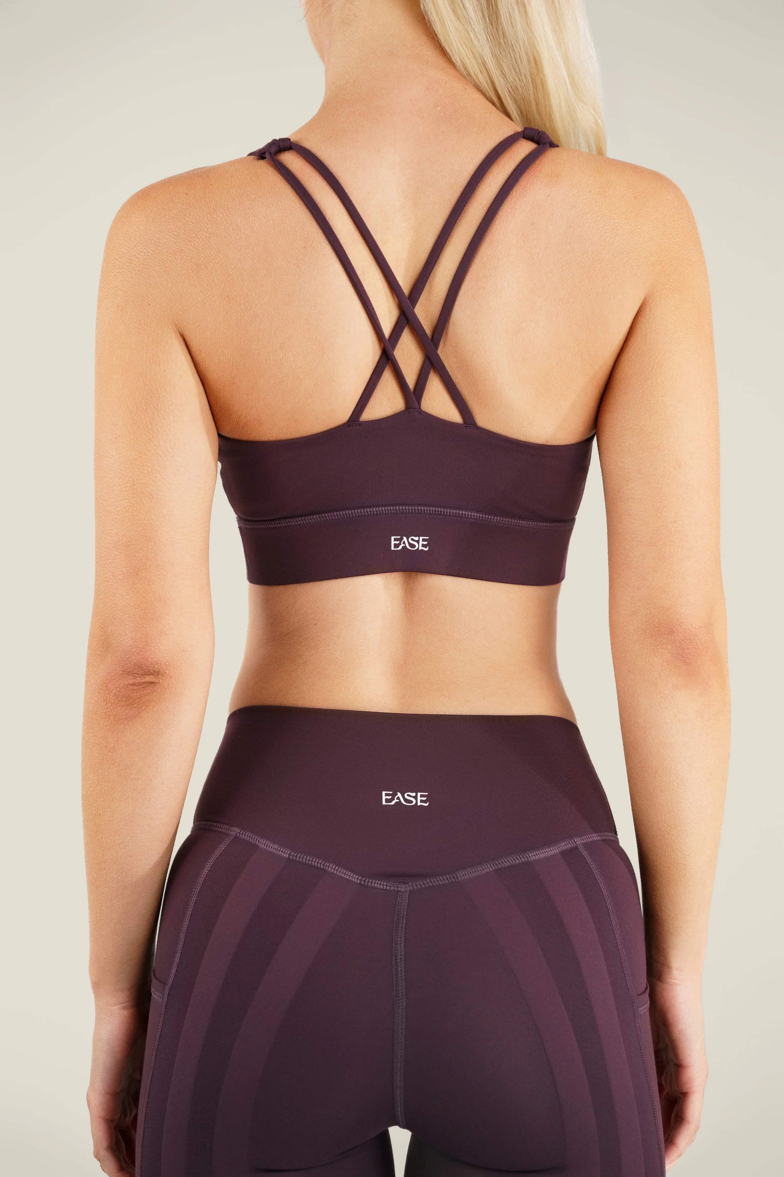 Back view of a woman, wearing a purple athletic sports bra with crisscross straps and high-waisted matching leggings, with the word 'EASE' printed on both the sports bra and leggings.