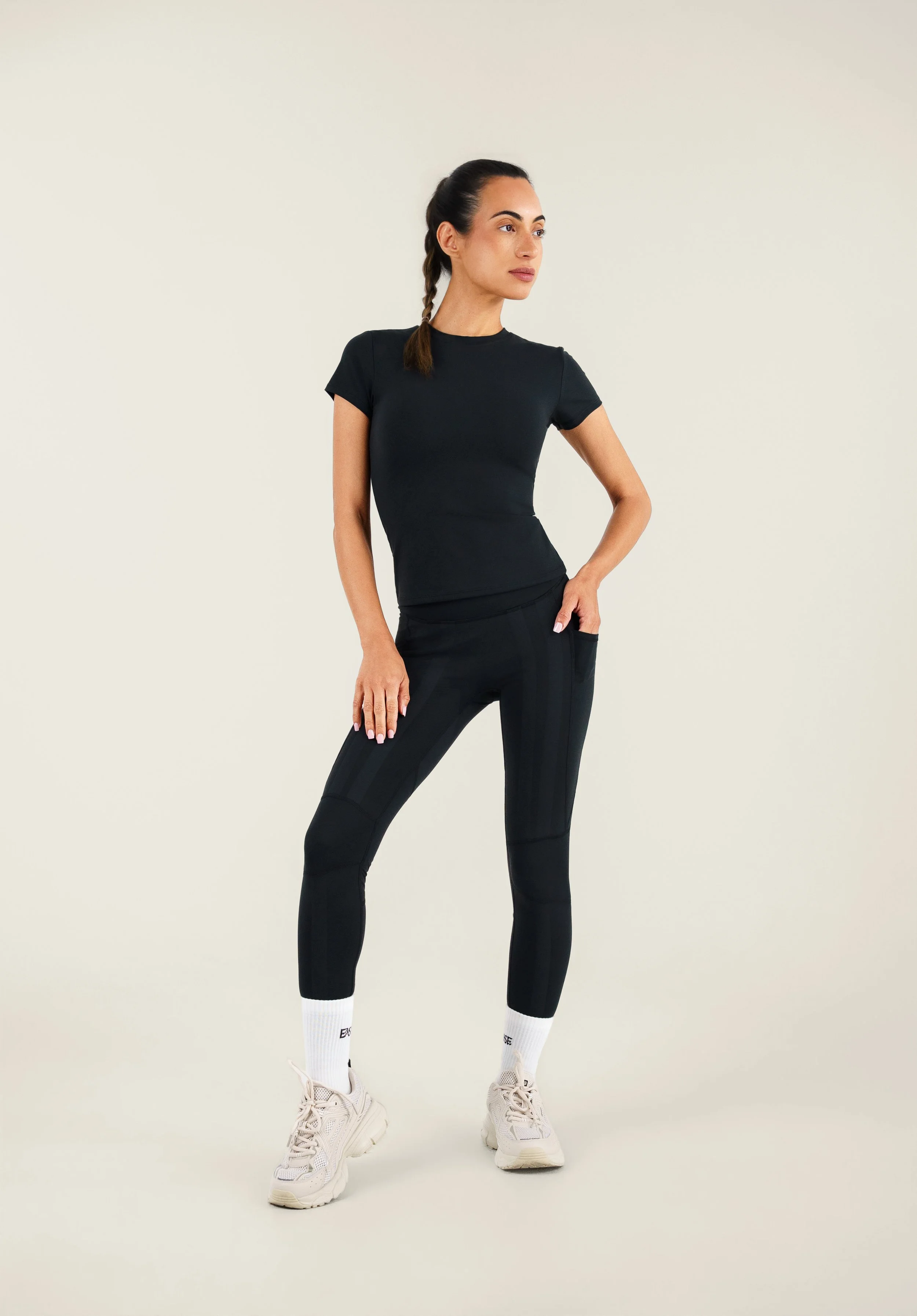Woman in black athletic wear standing with one hand on hip and looking to the side, against a plain light background.