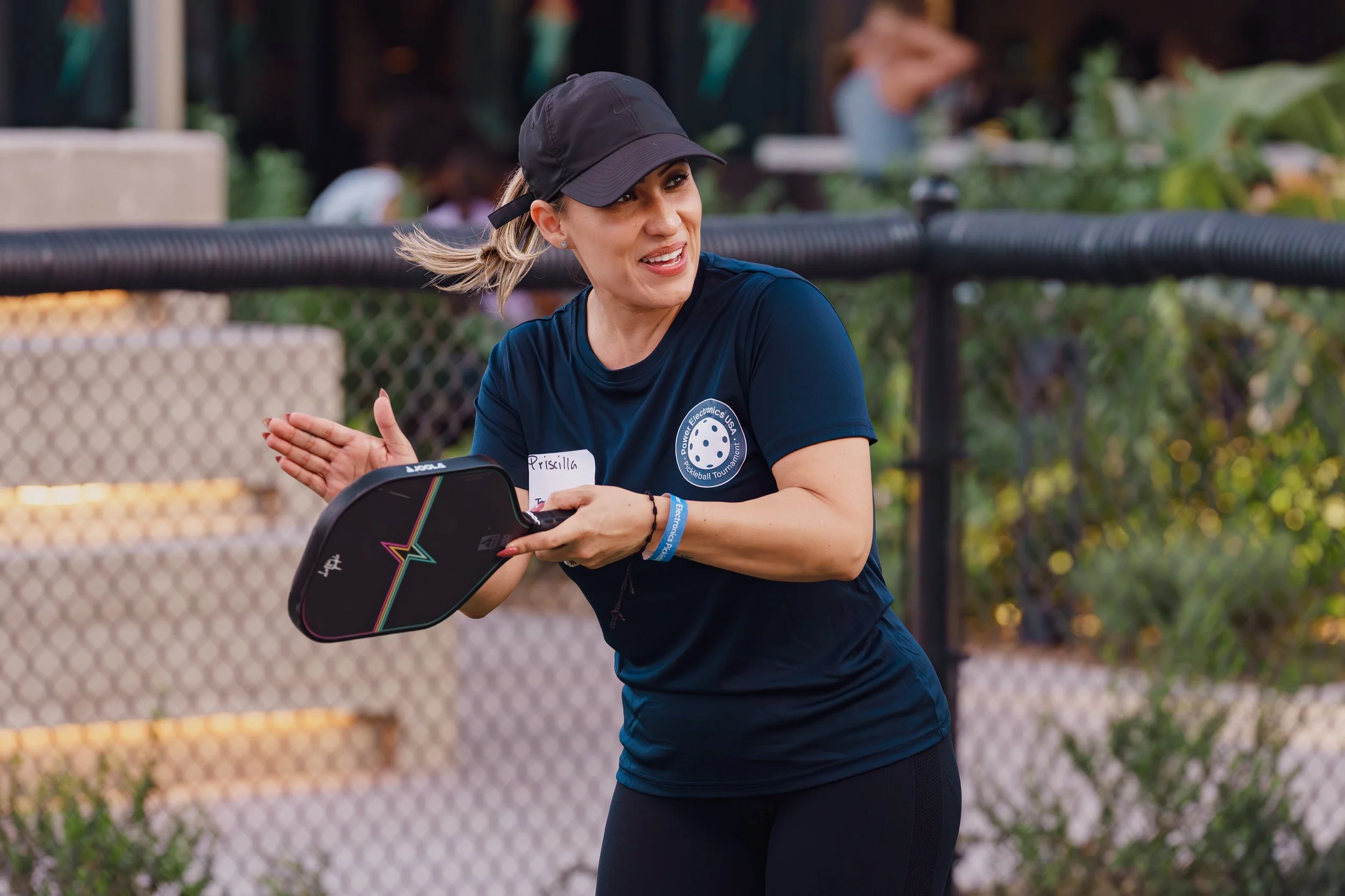 A woman with a black cap and a navy blue sports shirt holding a pickleball paddle, smiling and clapping, outdoors near a chain-link fence with greenery and other people in the background.