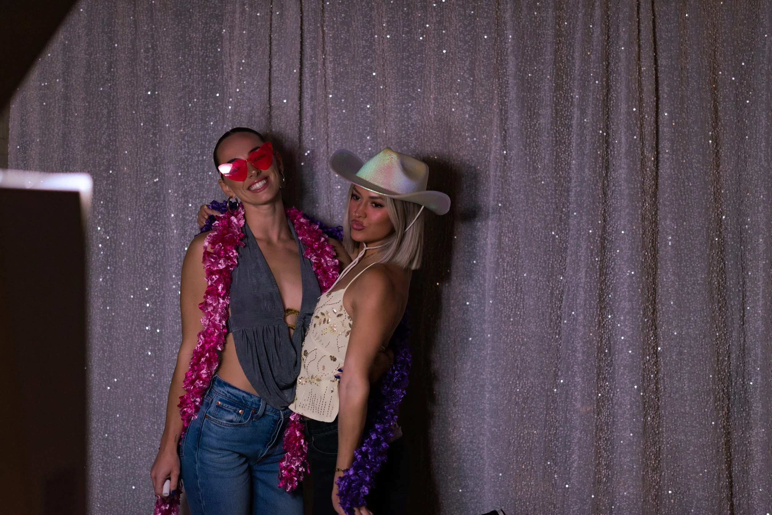 Two women standing together in front of a glittery curtain, smiling and posing for a photo. One is wearing heart-shaped sunglasses, a lei, a deep v-neck top, and jeans. The other is wearing a shiny cowboy hat, a sleeveless top, and a lei.
