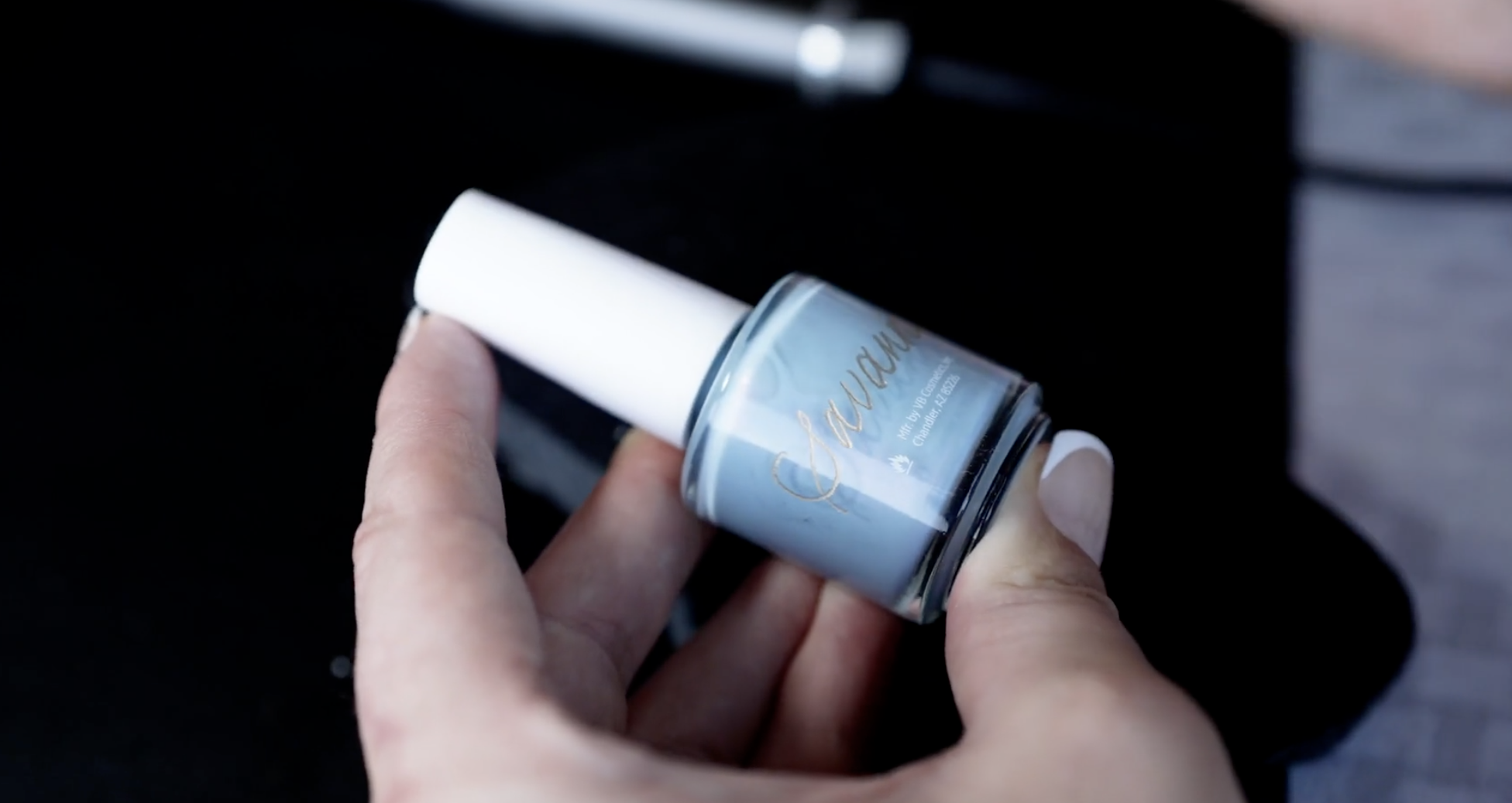 A person's hand holding a small jar of light blue nail polish with white cap, labeled with gold lettering.