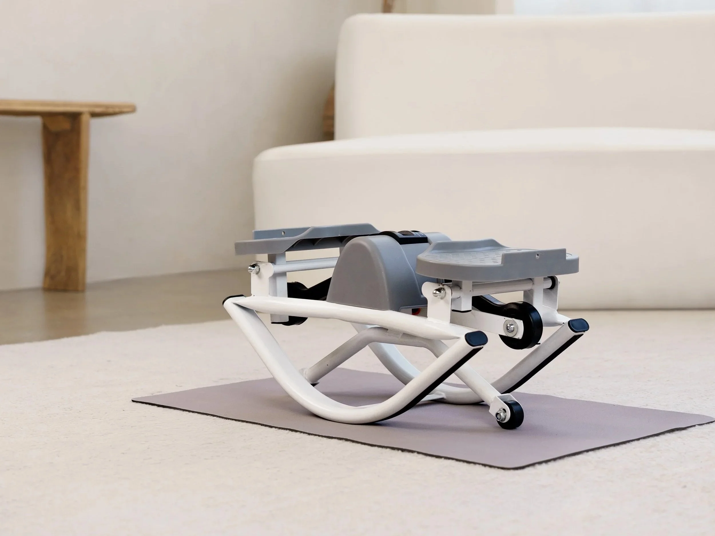 A gray stair stepper exercise machine placed on a beige carpet in a room with light-colored walls, next to a light-colored couch and a small wooden side table.