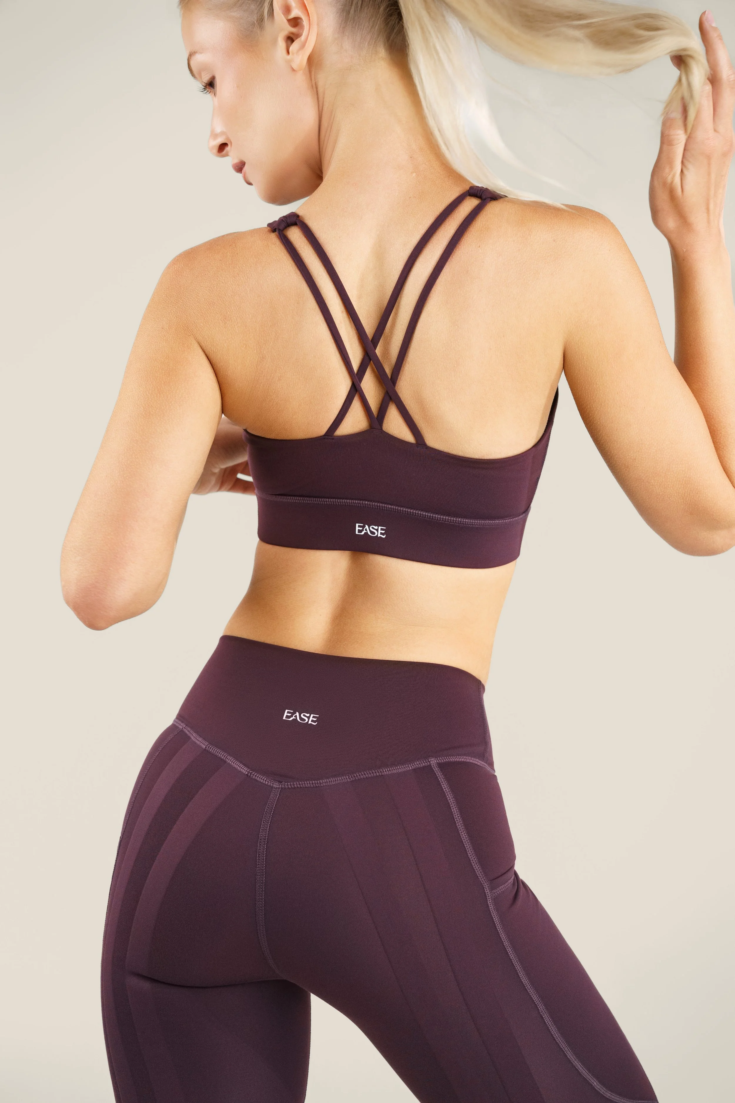 A woman wearing a purple sports bra and matching high-waisted leggings, with the word 'EASE' printed on both, is posing with her back to the camera, holding her hair with one hand against a plain light-colored background.