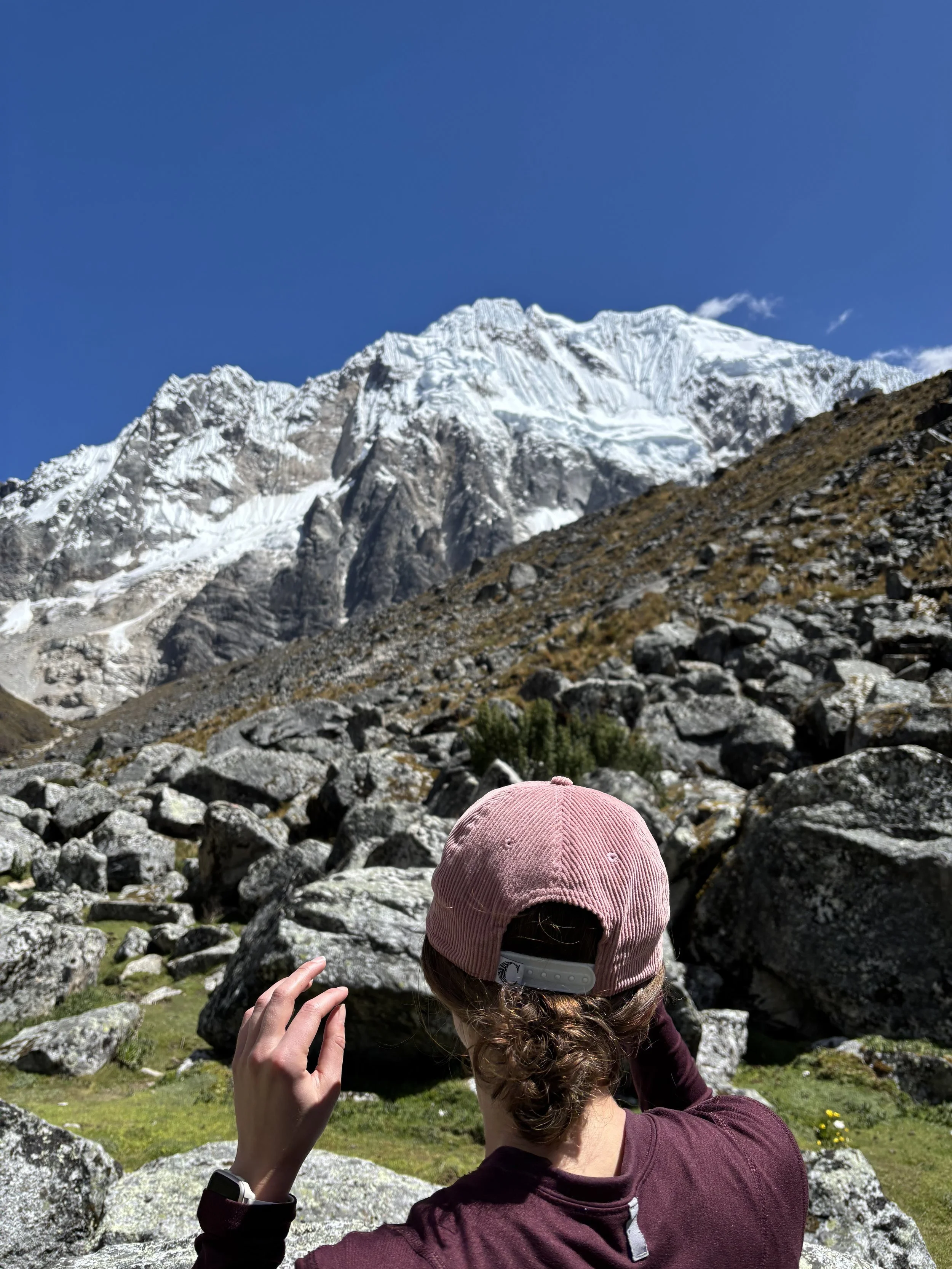 A woman wearing a pink North Carolina cap and maroon jacket standing on rocky terrain and taking a photo of a snow-capped mountain under a clear blue sky.