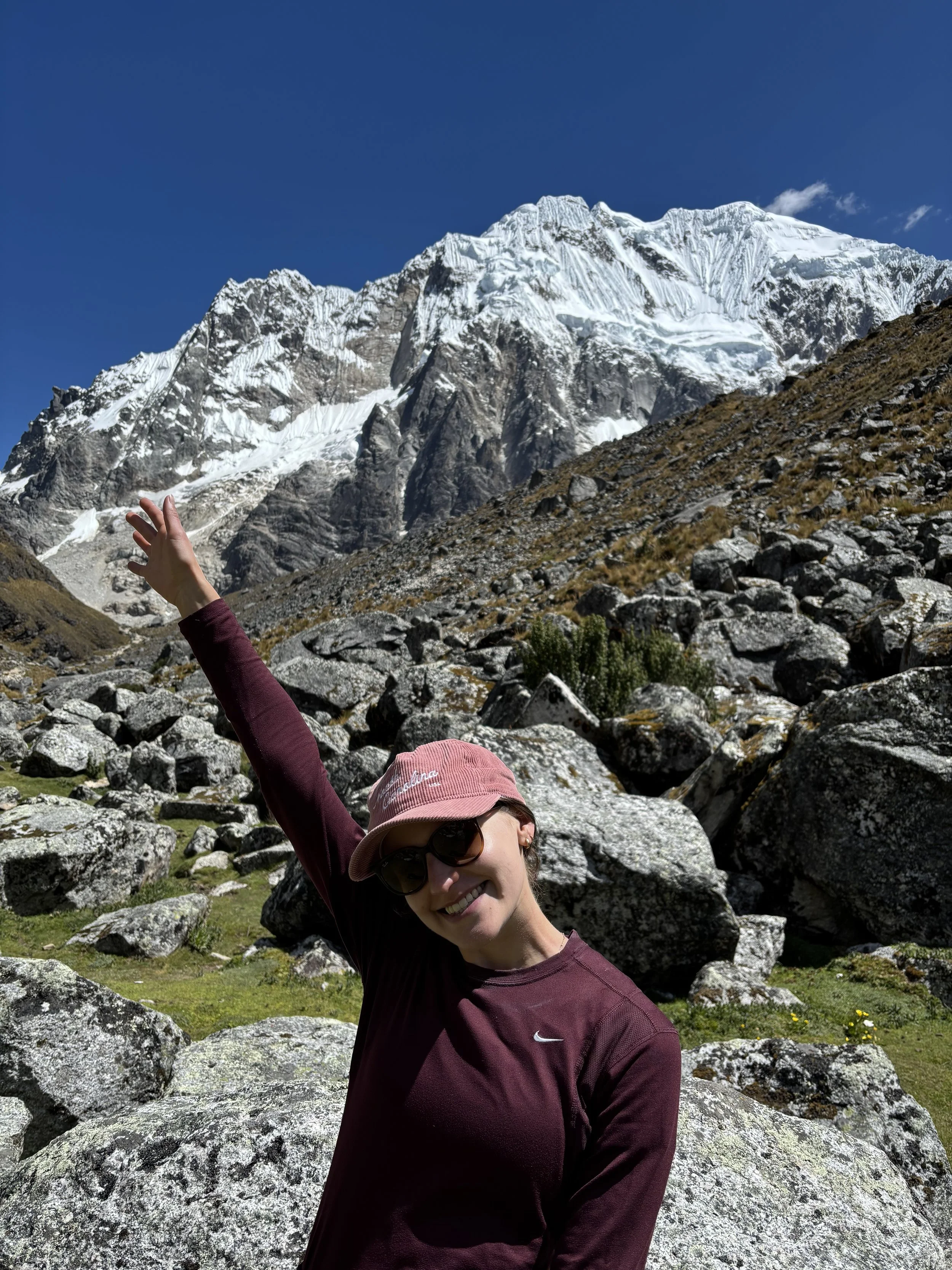 A woman smiling and wearing sunglasses, a pink North Carolina corduroy hat, and a maroon shirt, is standing among rocks with a mountain range featuring snow-capped peaks in the background.
