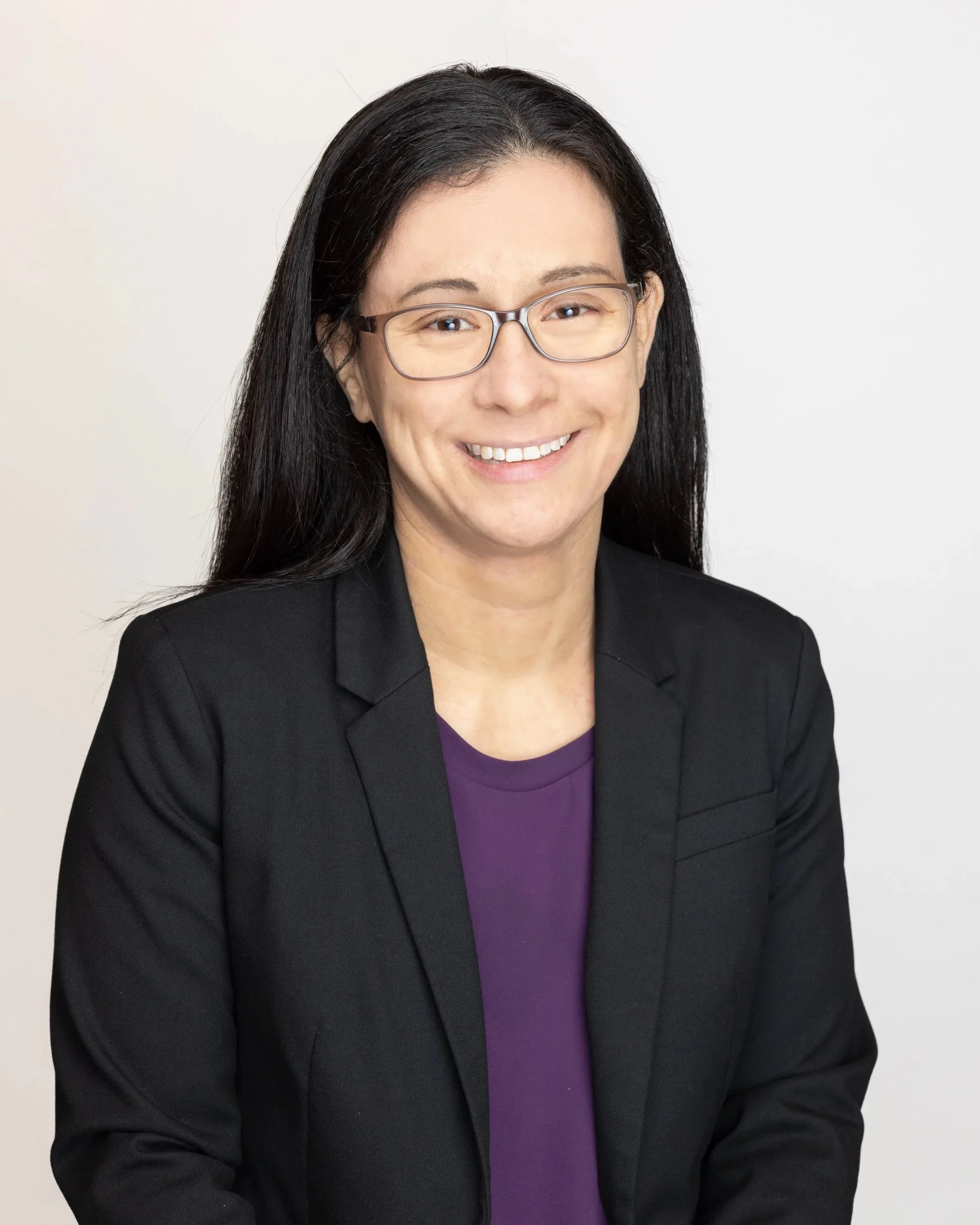 Person with long black hair, wearing glasses, a purple shirt, and a black blazer, smiling against a plain background.