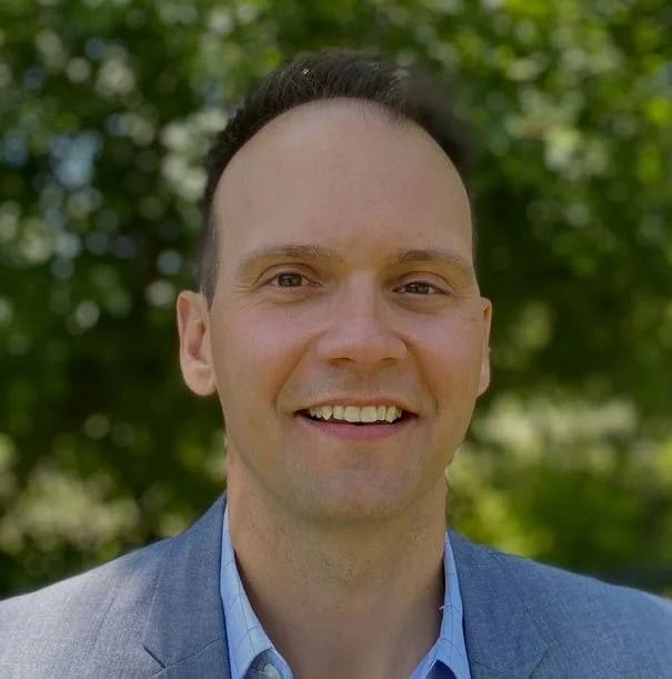 A man with glasses and a beard smiling, wearing a black shirt against a gray background.