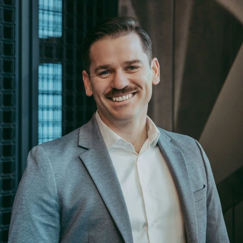 Man in a black suit jacket and white shirt smiling against a white background.