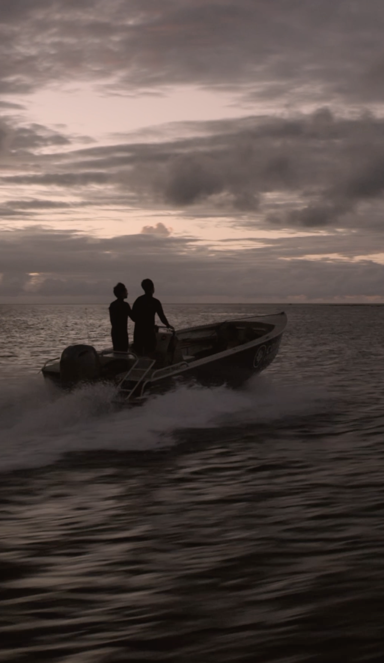 Two people on a boat during sunset or dusk, silhouetted against the sky with clouds, moving across the water.