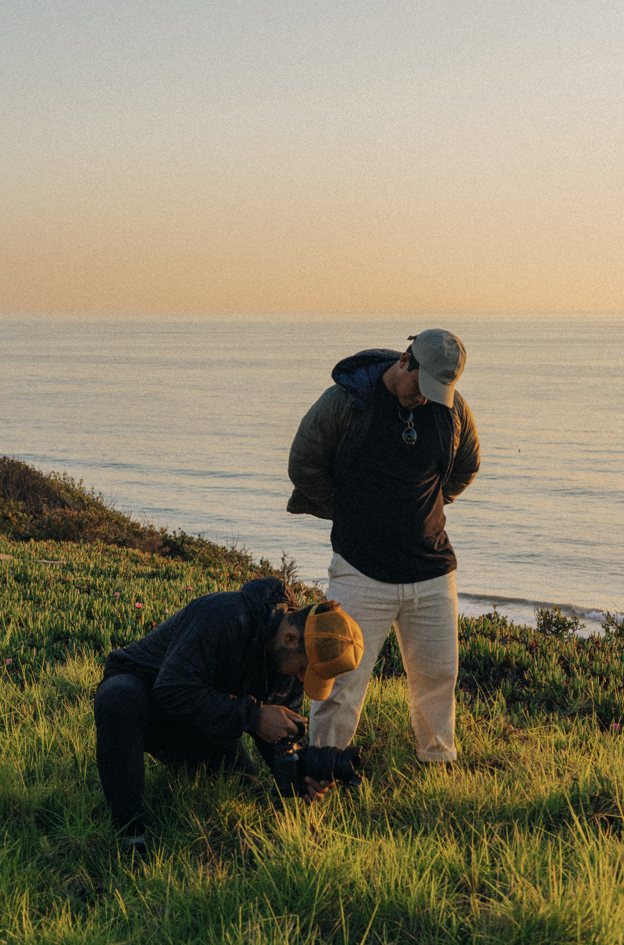 Two people by the ocean during sunset; one is sitting and taking a photo with a camera, the other is standing observing, both wearing jackets and caps, surrounded by green grass and coastal vegetation.