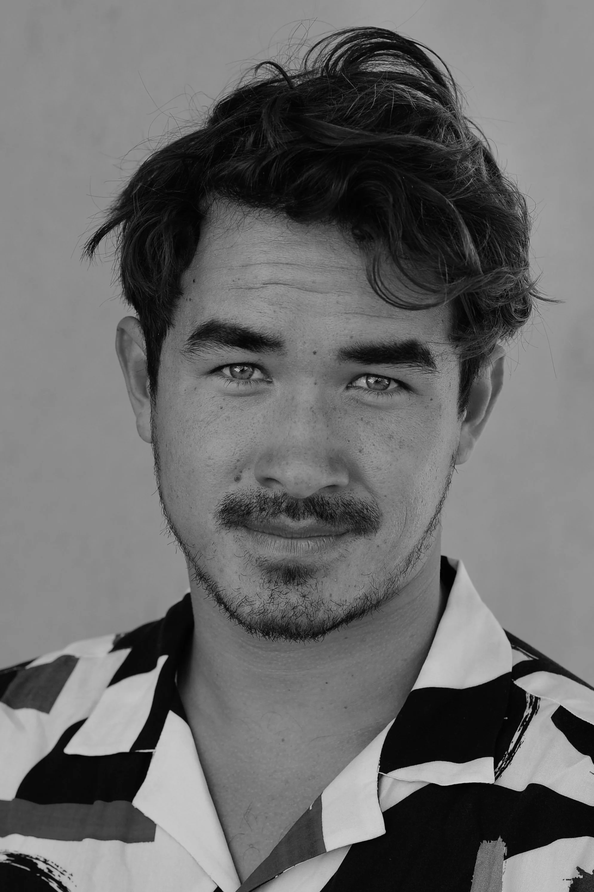 Black and white portrait of a young man with dark, wavy hair, a trimmed beard, wearing a patterned shirt.