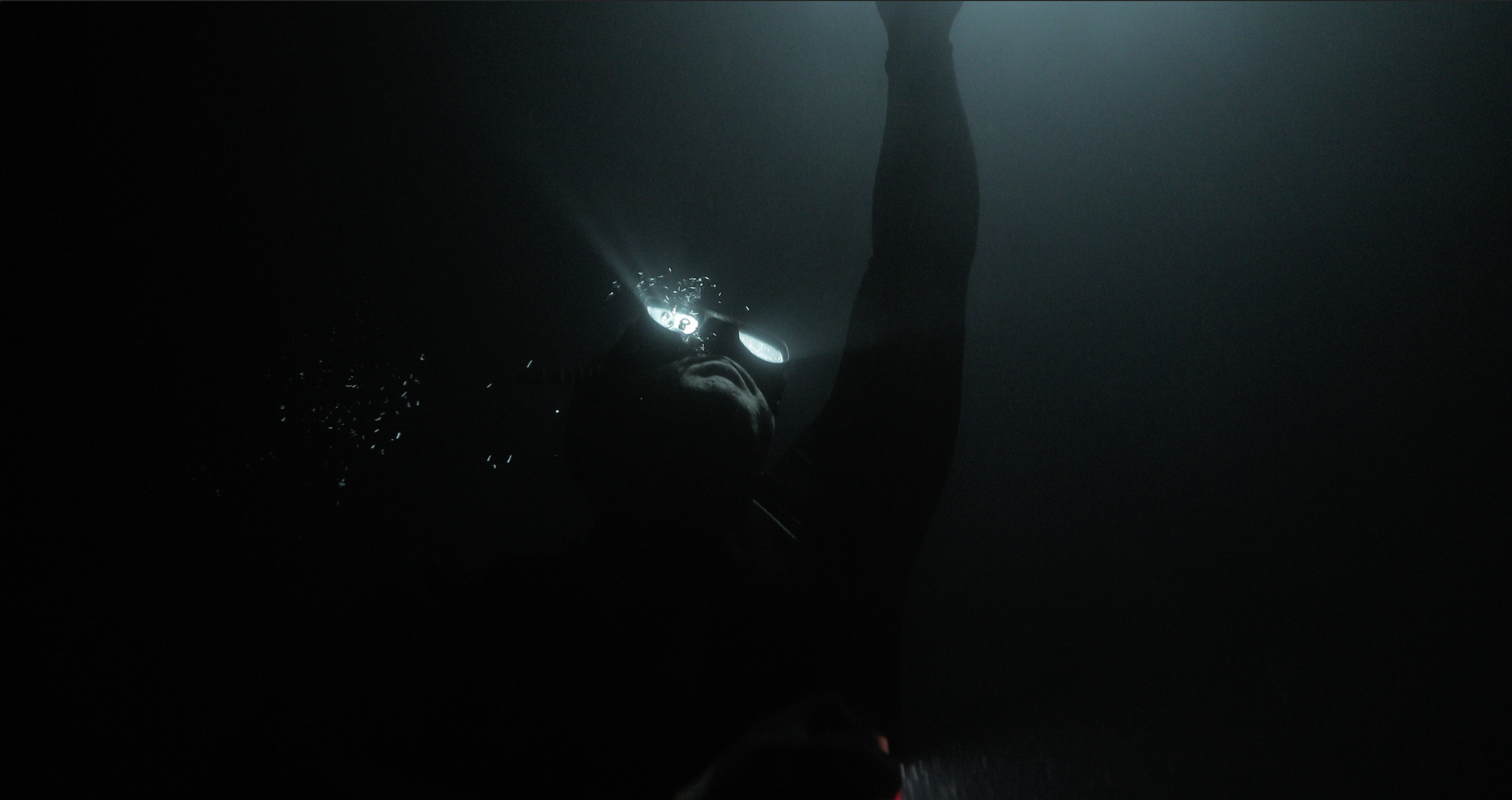 Person with a snorkel and mask swimming underwater, illuminated by a flashlight, in a dark environment.