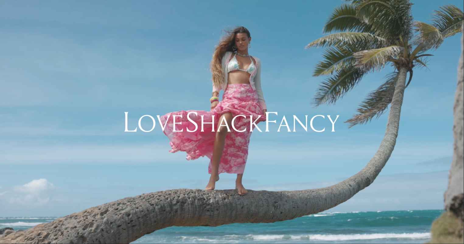 A woman standing on a curved palm tree at the beach, wearing a bikini top and a pink skirt, with the ocean and a blue sky in the background, and the words "LoveShackFancy" overlaid.