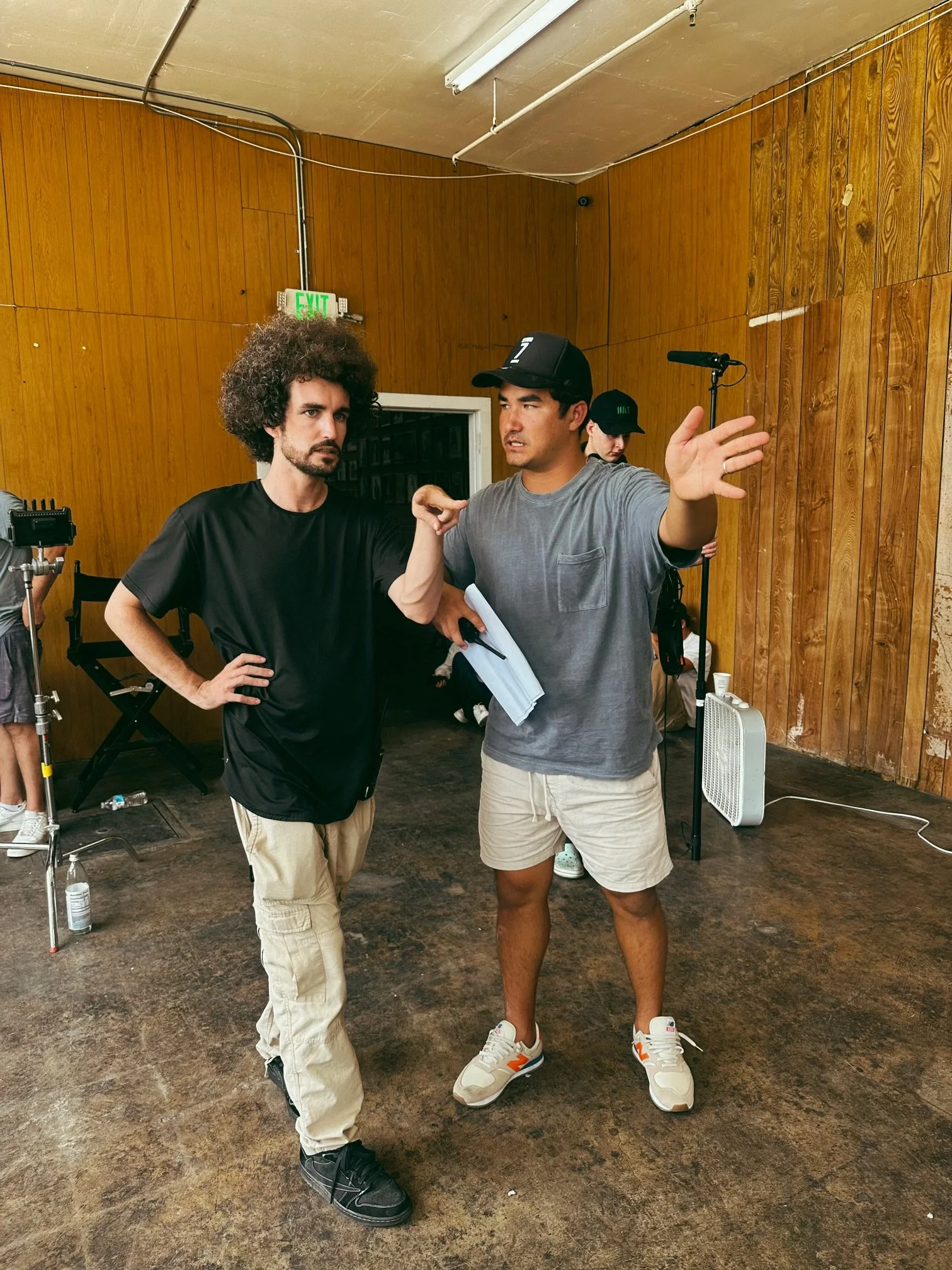 Two men are standing in a room with wood-paneled walls, apparently engaged in a conversation or discussion. One man with curly hair in a black shirt has a serious expression, while the other man in a gray t-shirt and baseball cap is talking and gesturing with his hands. Equipment like microphones, a fan, and other objects are visible in the background, indicating this might be a film or TV set.