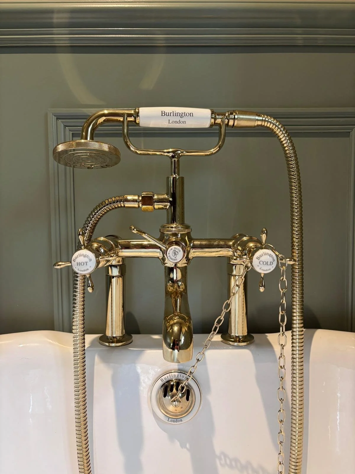 A vintage, gold-colored faucet with separate hot and cold water controls, a shower head attachment, and a chain for a plug, all mounted above a white sink. Labels read 'Burlington London', 'HOT', and 'COLD'.