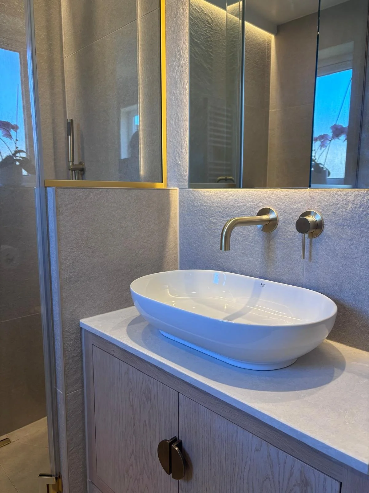 Modern bathroom with a white oval vessel sink on a light wooden cabinet, gold wall-mounted faucet, textured beige wall, large mirror, and a glass-enclosed shower with a built-in shelf and a window with a potted plant outside.