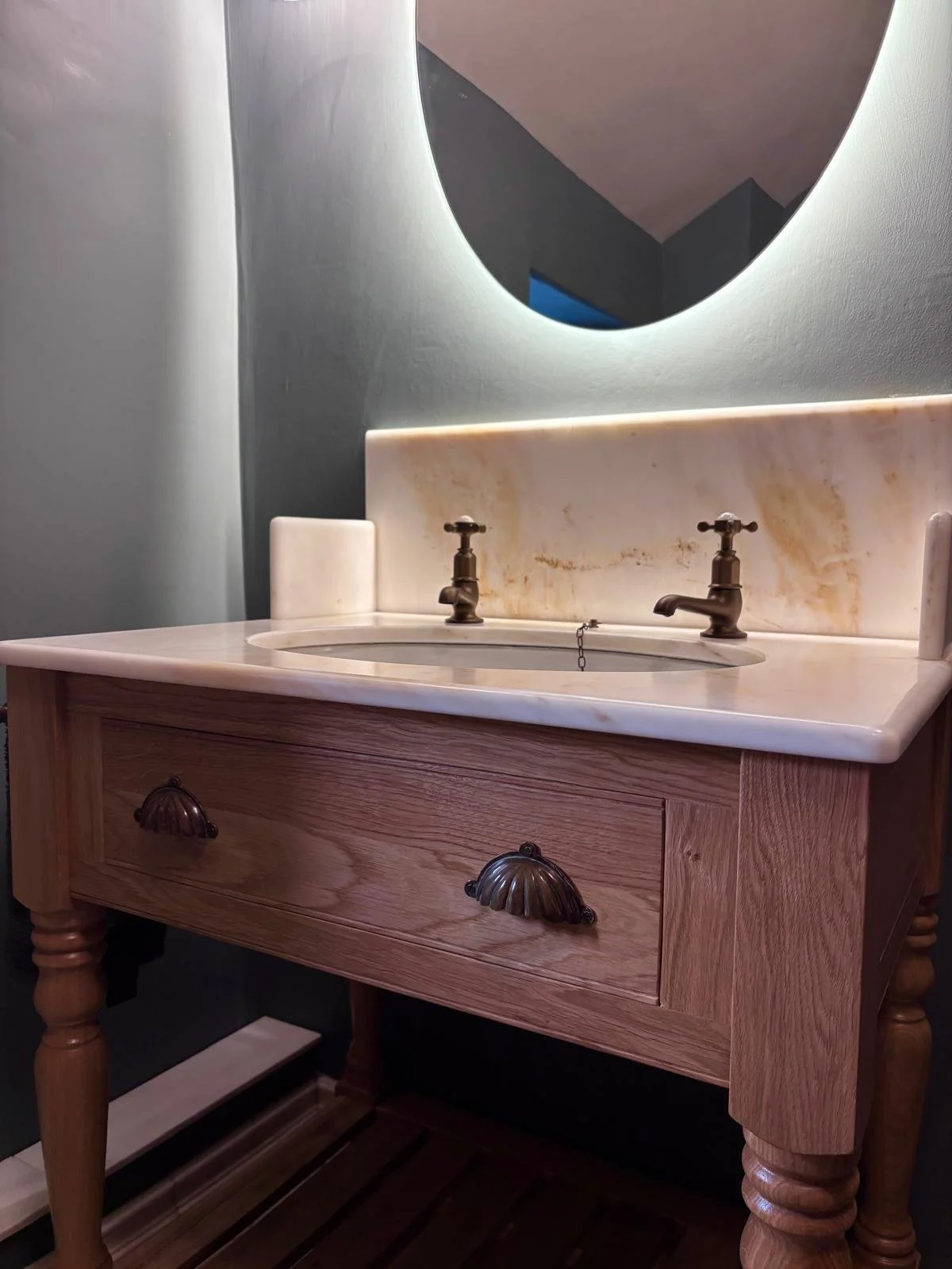 A vintage wooden bathroom vanity with a marble top, two bronze faucets, and a large oval mirror with backlit lighting.