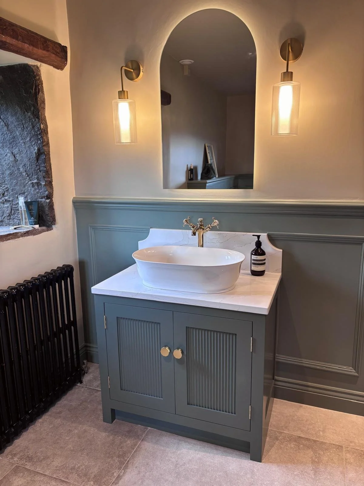 A bathroom vanity with a white vessel sink, gold-tone faucet, dark green cabinet with beadboard doors, and a marble countertop. Two wall-mounted light fixtures with cylindrical glass shades, a mirror with an arched top, and a small window with stone trim are visible.