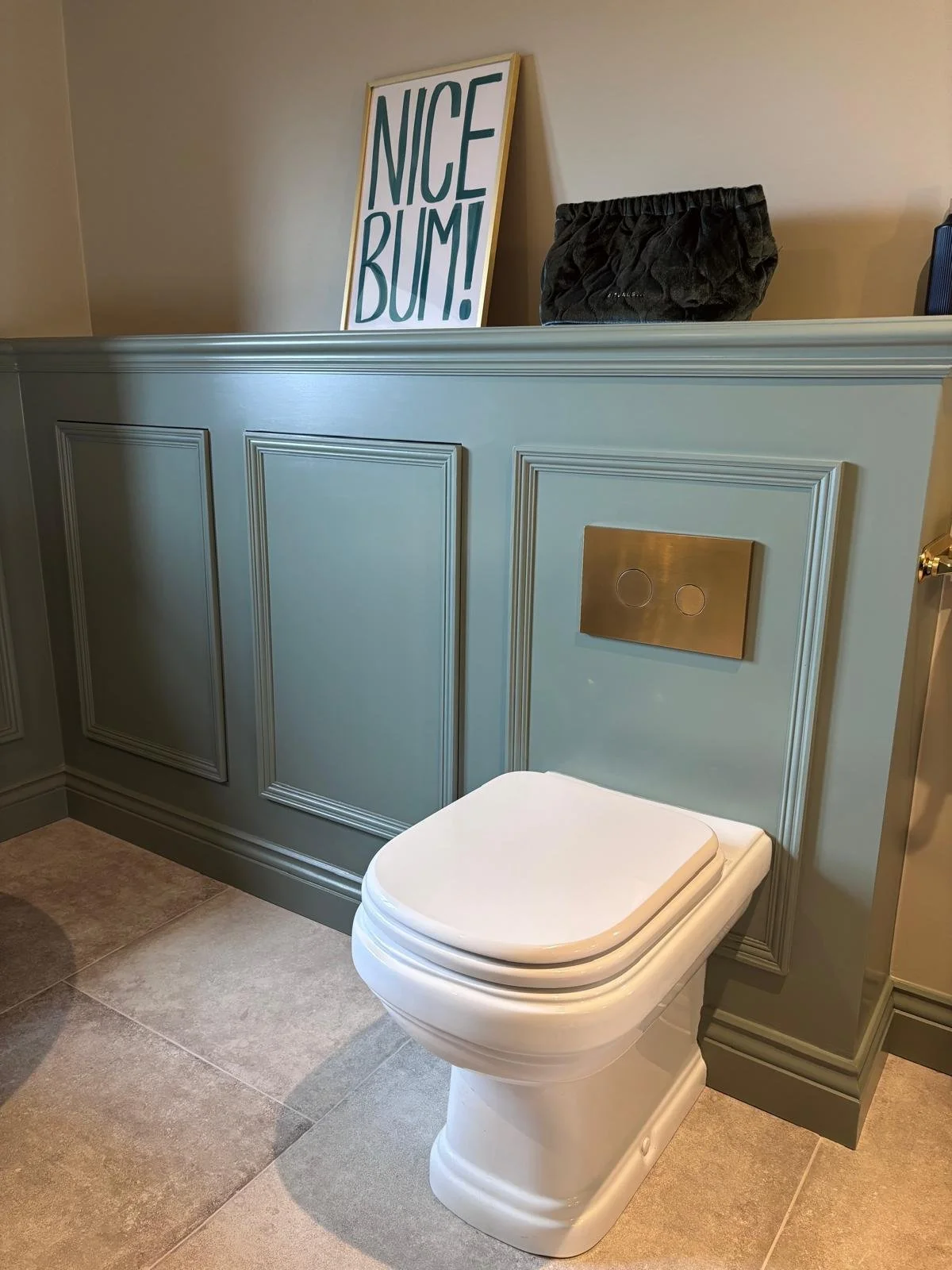 A modern toilet in a bathroom with light beige tiled floor and decorative light blue wall paneling. There is a framed sign saying "NICE BUM!" leaning against the wall and a black textured bag placed above the toilet.