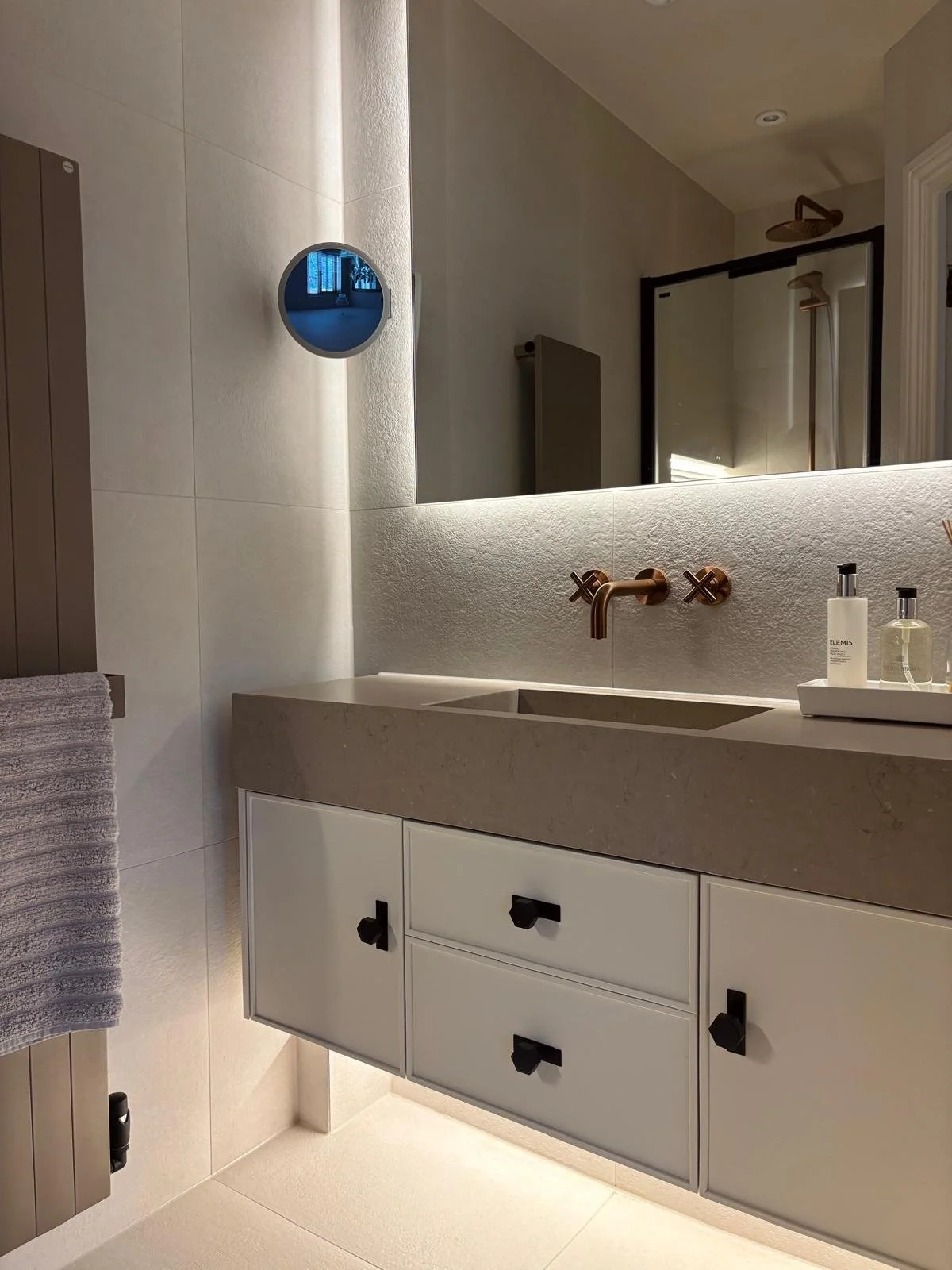Modern bathroom with beige countertop, copper faucet, white cabinet with black knobs, large mirror, soap dispensers and bottles, textured wall, towel hanging on the left, shower with glass door and copper showerhead in the background.