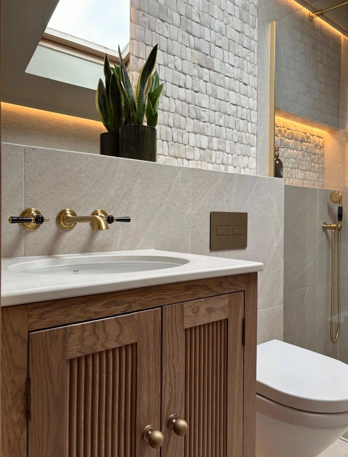 A modern bathroom with a wooden cabinet, a white sink, gold fixtures, a plant on a backsplash, and a white textured wall with warm lighting.