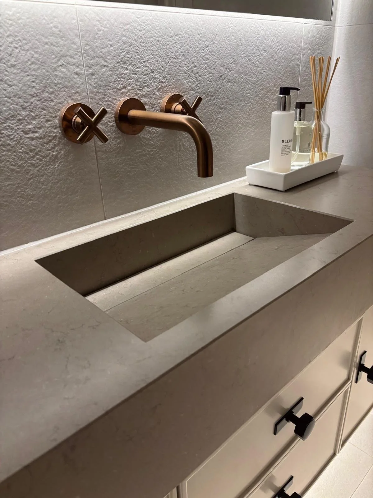 Bathroom sink with a copper faucet and two cross handles, with bottles of soap and diffuser sticks on a white tray nearby.