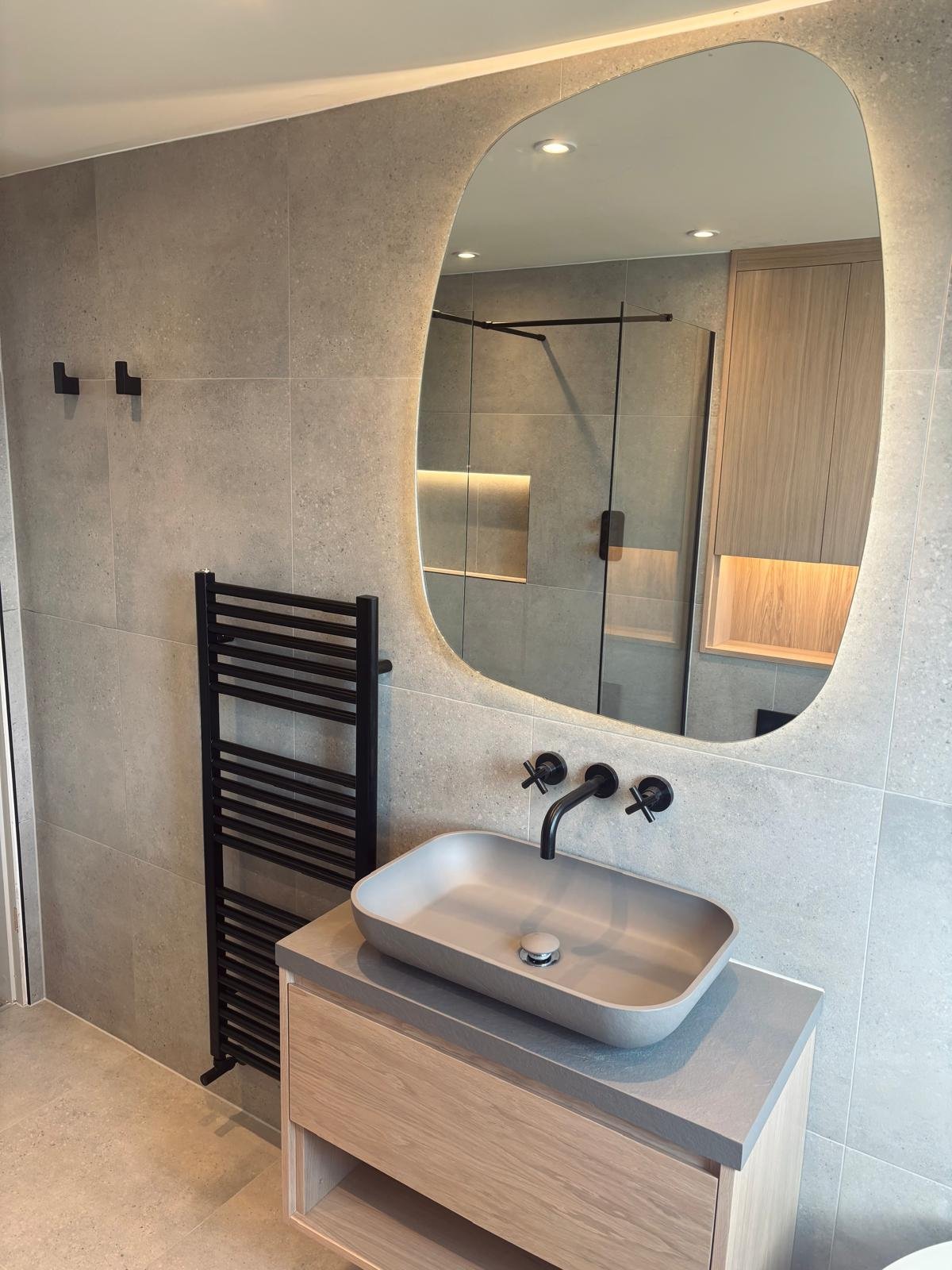 Modern bathroom with a rectangular vessel sink on a wooden vanity, black wall-mounted faucet, large mirror with rounded edges, black heated towel rack, and neutral-toned tiled walls and floors.