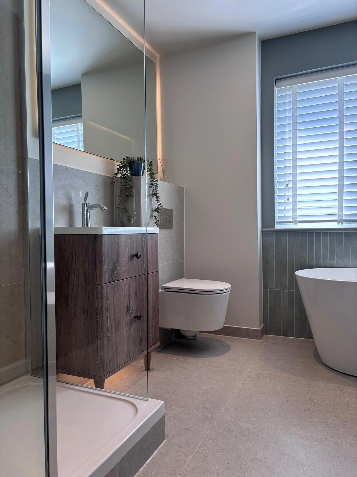 Modern bathroom with a glass shower, wooden vanity, white toilet, freestanding bathtub, window with white blinds, and neutral tones.