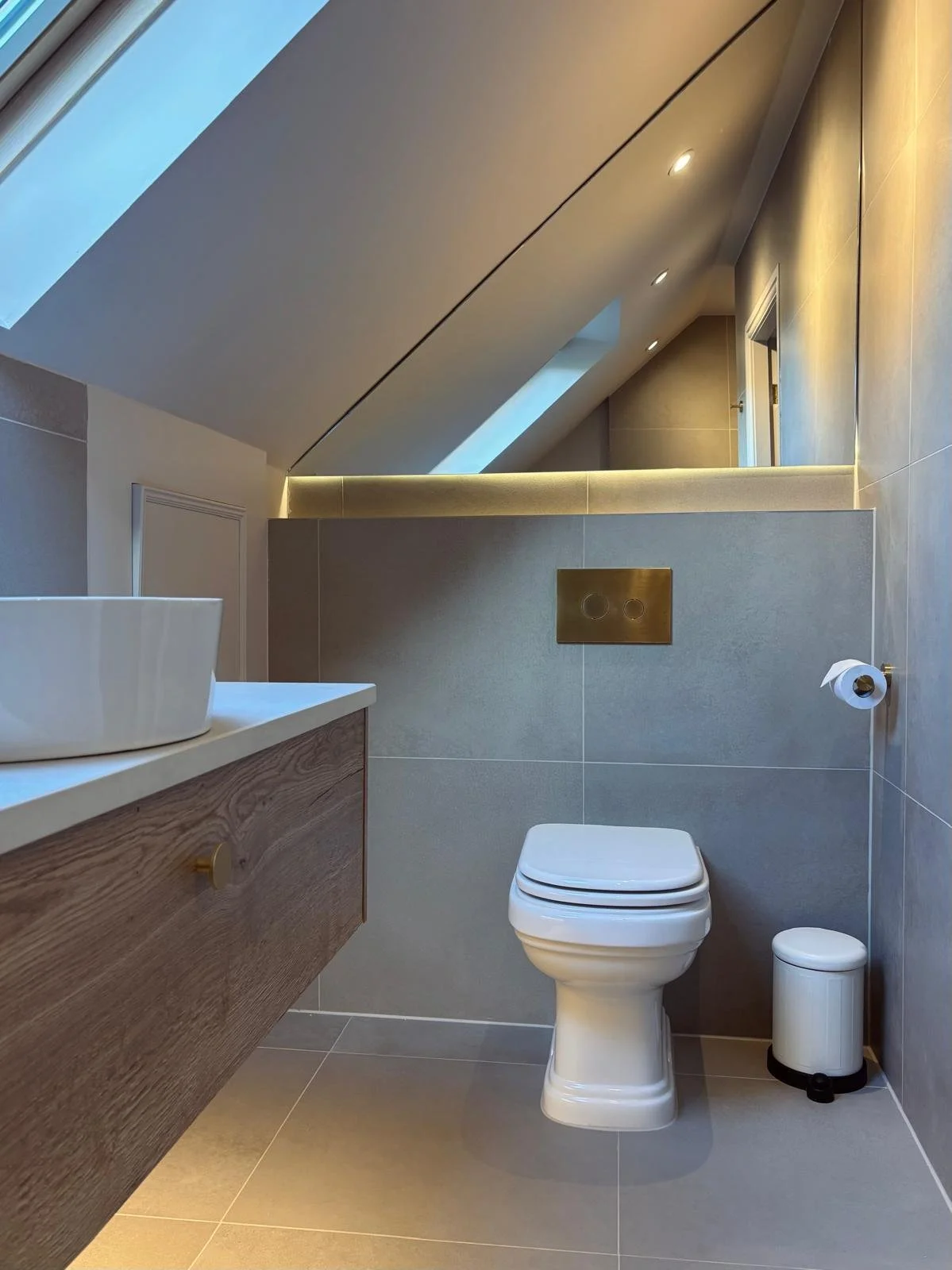 Modern bathroom with gray tiled walls, a white toilet, a small wooden vanity with a white vessel sink, a toilet paper roll, and a trash bin. The ceiling features angled design with skylights and recessed lighting.